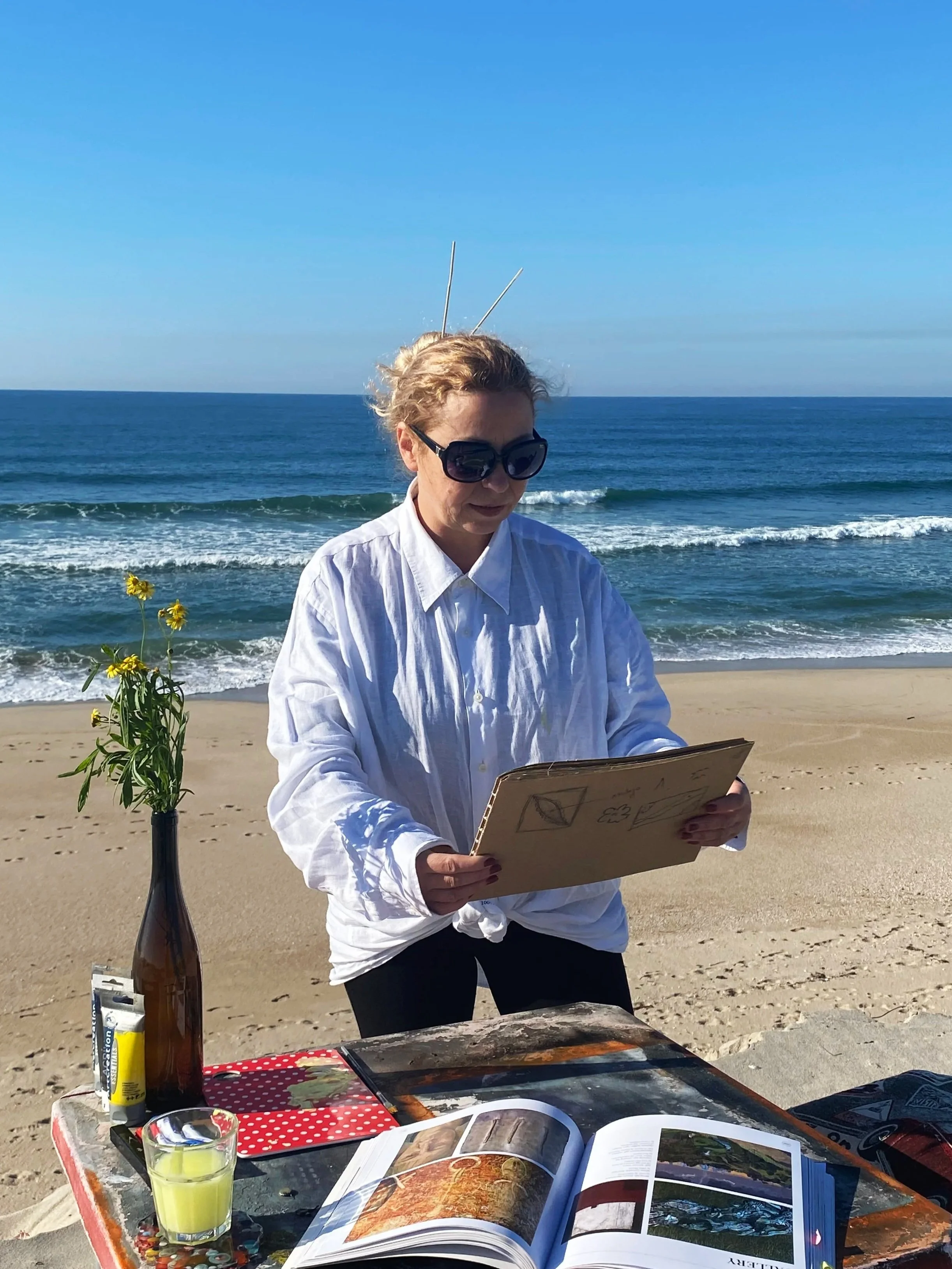 Pessoa com cabelo claro e óculos escuros observa um quadro na praia, enquanto uma mesa com livros, um copo de suco, uma garrafa de tinta, molde de pintura e material de arte está na frente.