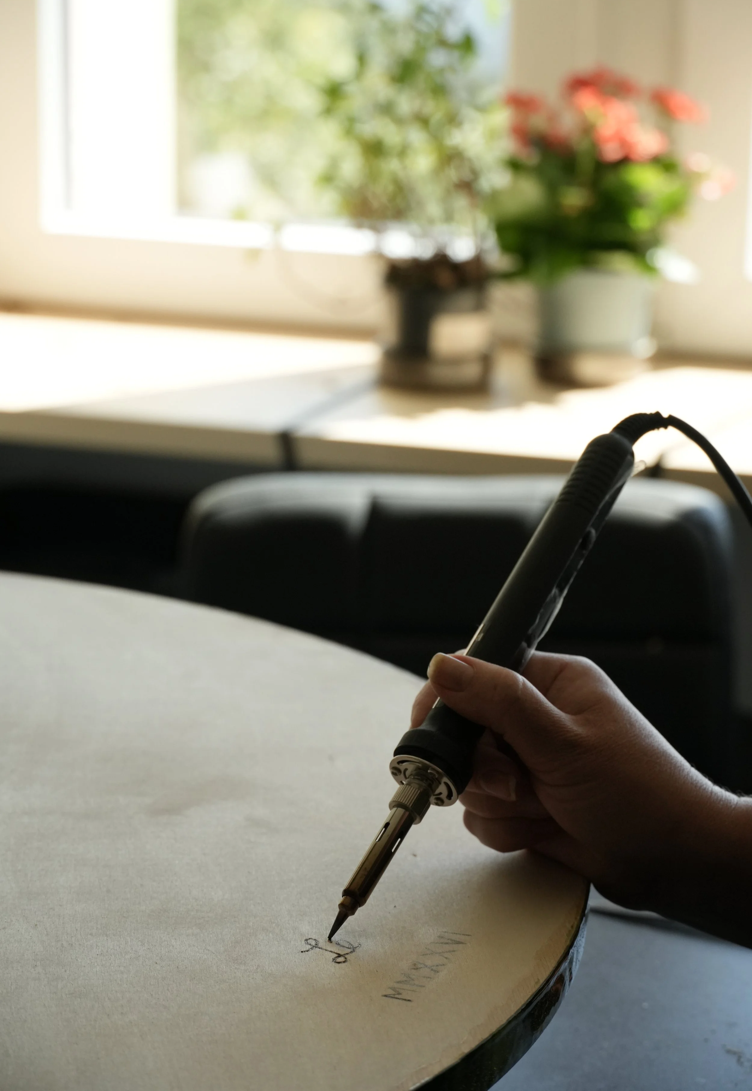 Pessoa escrevendo no caderno com uma caneta ponta fina em uma mesa, ao fundo uma janela com plantas.
