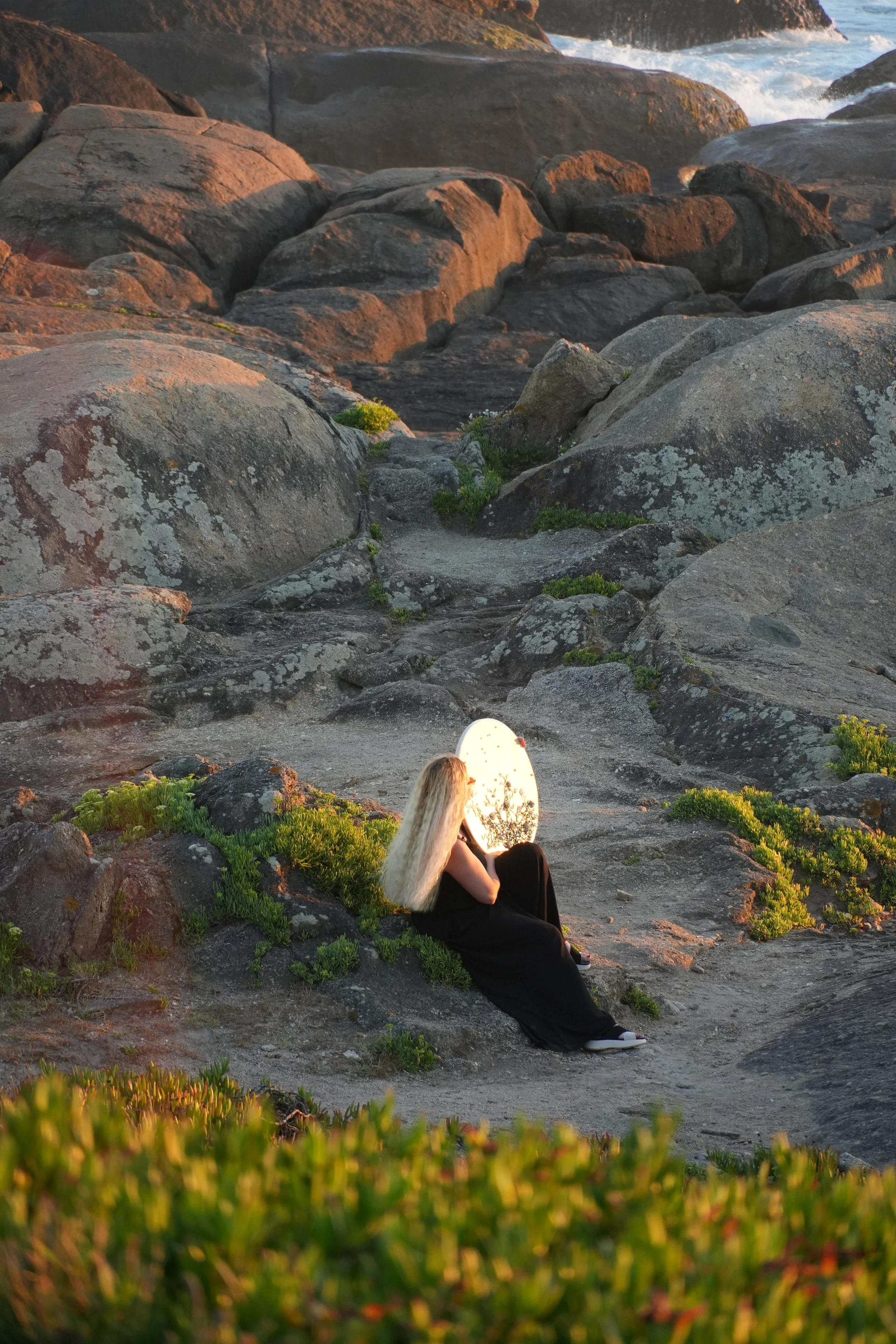 Lé sentada na areia da praia, segurando a nova obra de arte ao pôr do sol.