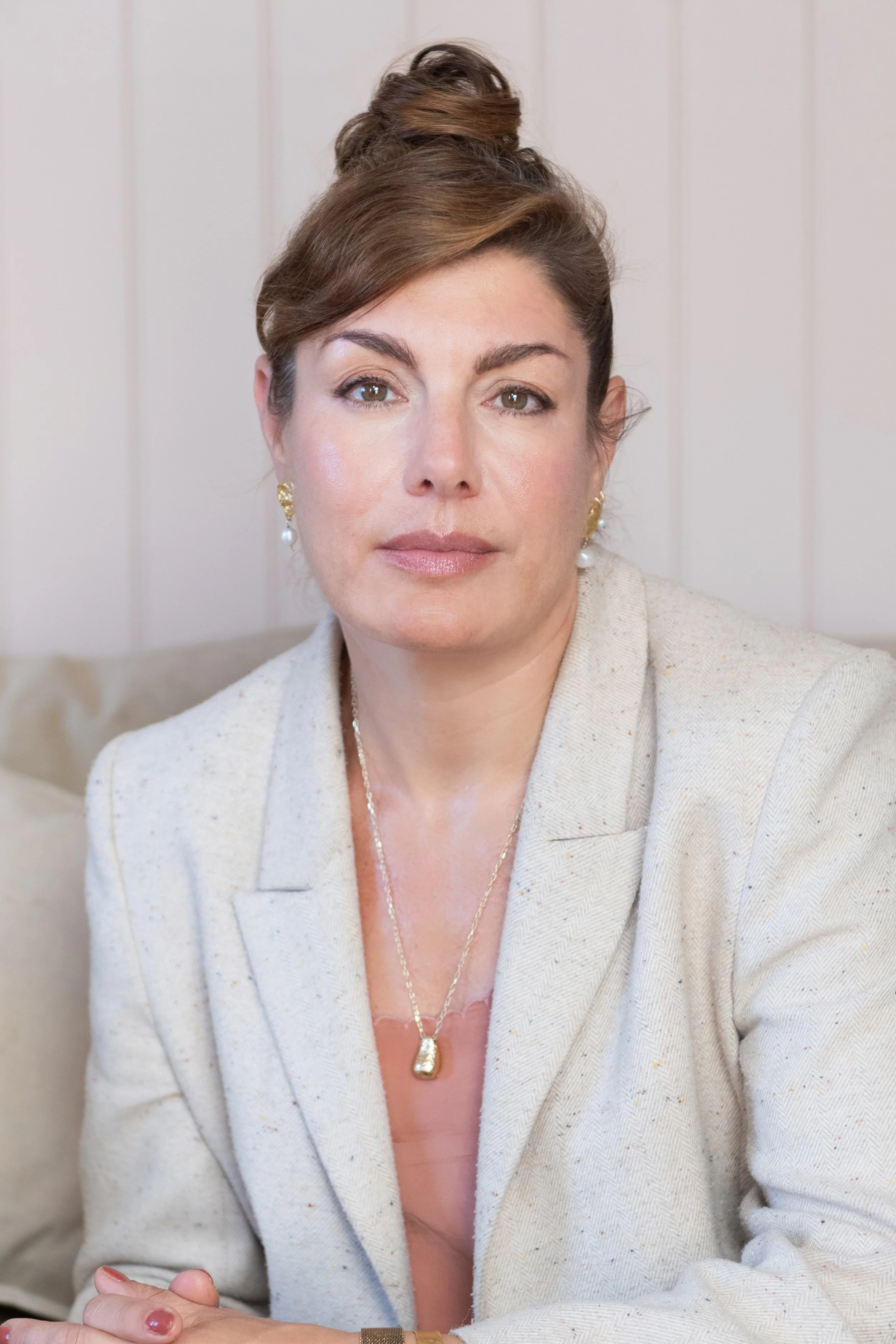 A woman with brown hair styled in a bun, wearing a cream-colored blazer and gold jewelry, sitting against a light-colored wood-paneled wall.