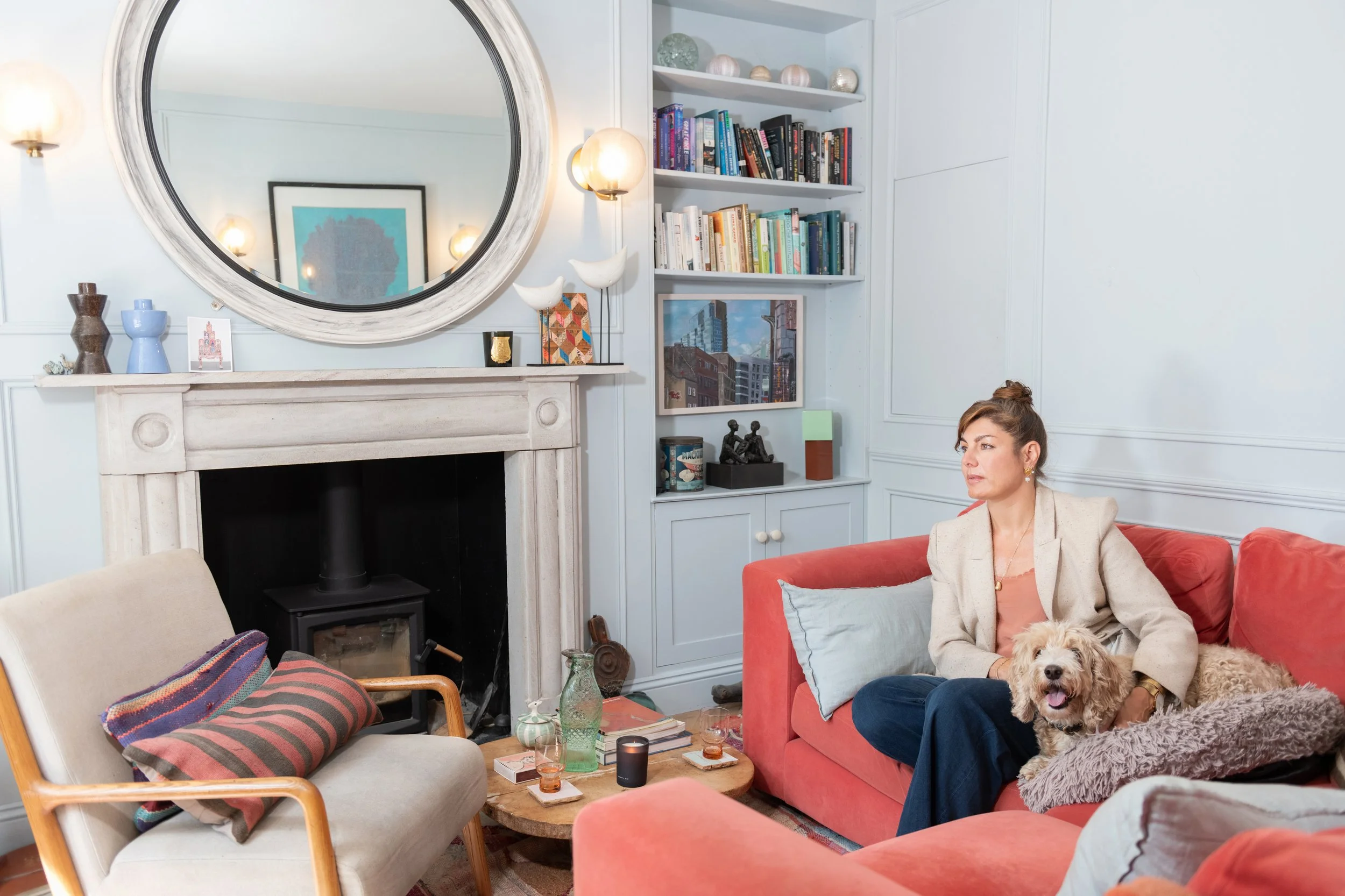 A woman sitting on a red couch with a dog on her lap in a cozy living room. The room has a large mirror above the fireplace, a bookshelf filled with books, and various decorative objects.