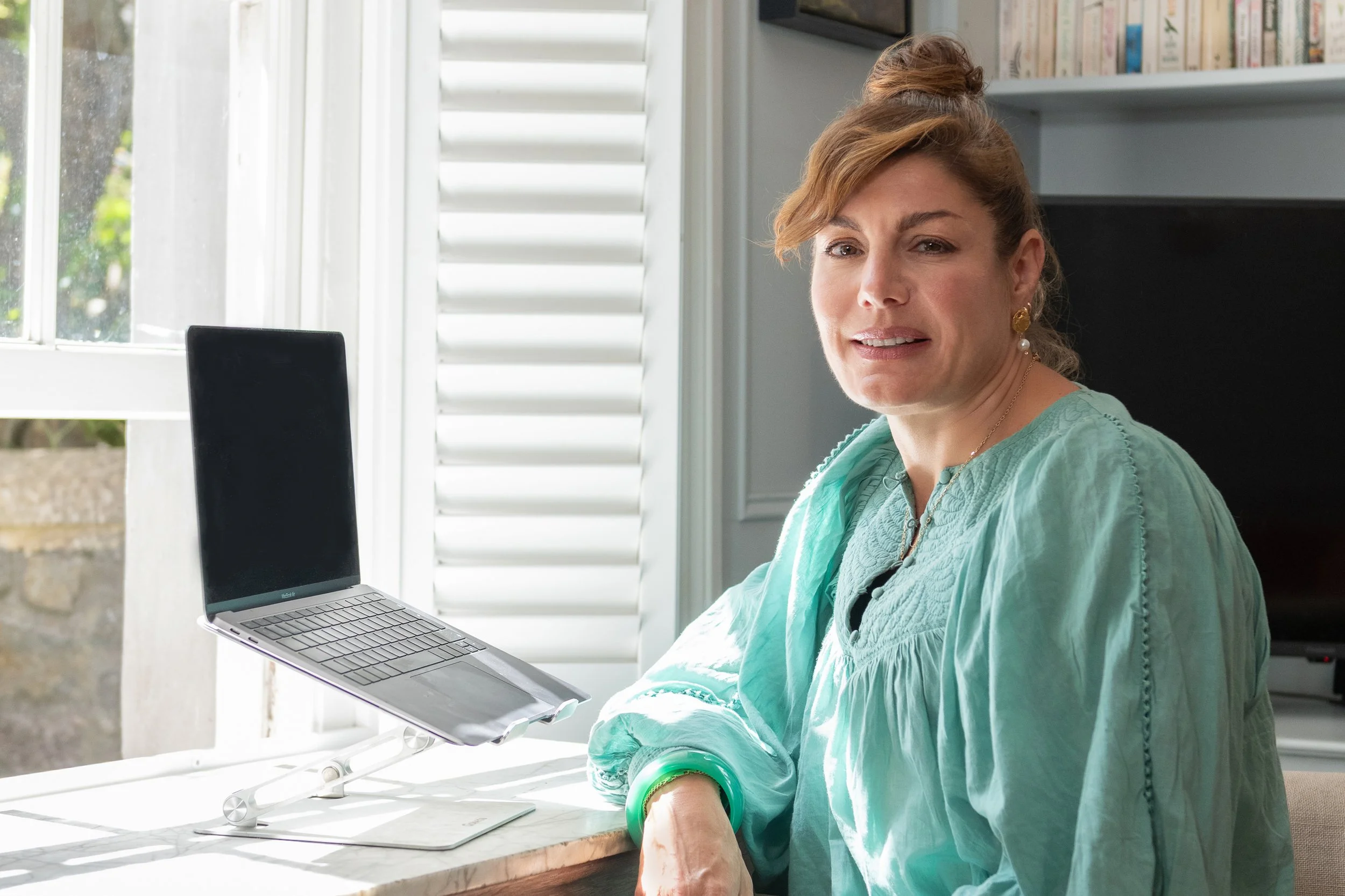 A woman with a top bun hairstyle sitting at a desk with a laptop in front of a window with white shutters, wearing gold earrings and a turquoise top.