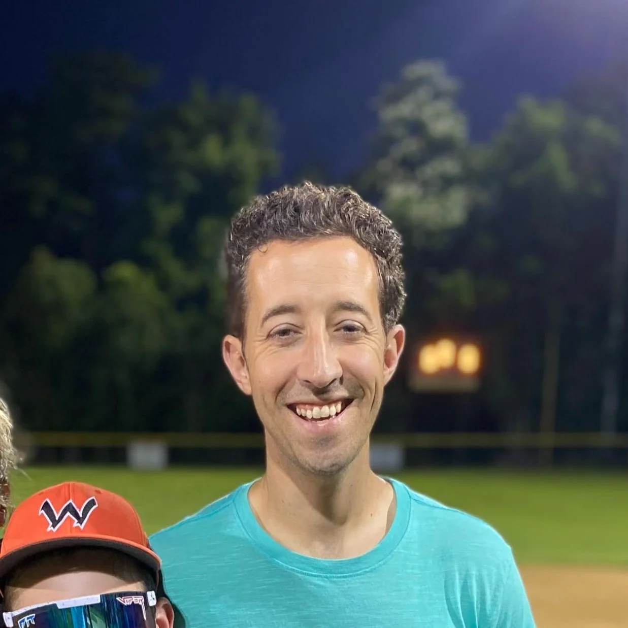 A smiling man standing outdoors at night, wearing a light blue T-shirt, with trees and a football field in the background.