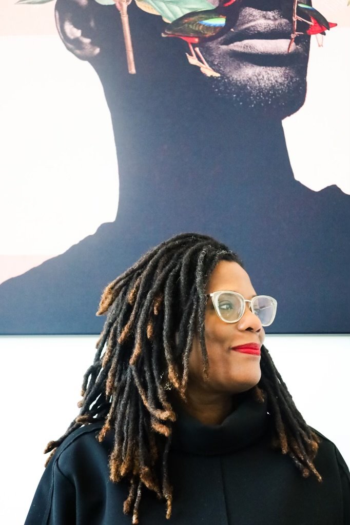 A woman with long dreadlocks, wearing glasses and bright red lipstick, standing in front of a large art piece depicting a person holding a fish.