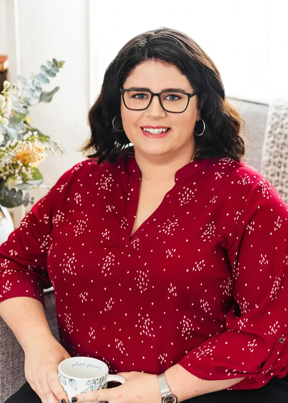 A photo of Linda Baggett, a therapist who is weight inclusive and HAES aligned. Linda is plus size, white, wears glasses and a red and white top. She is sitting with flowers behind her. 90094 | 90277 | 90402