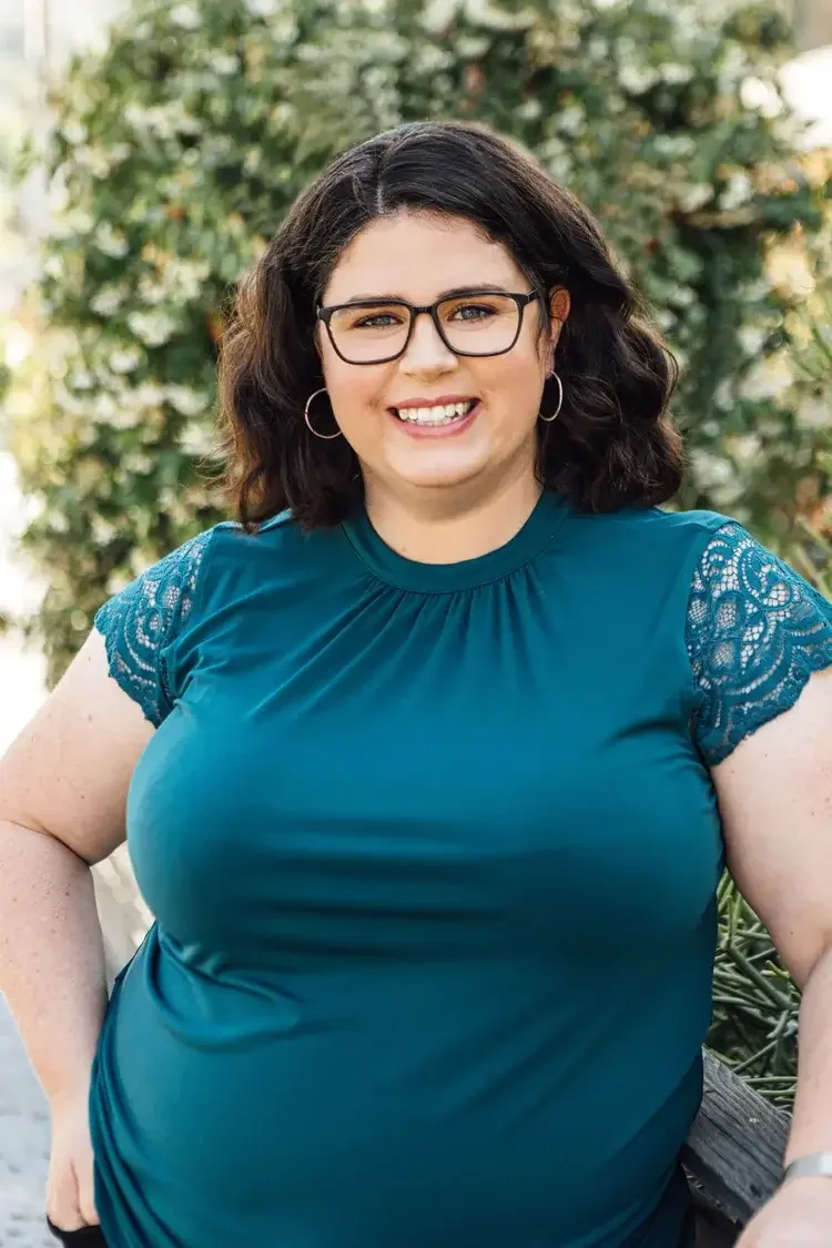 Headshot of Dr. Linda Bagget wearing a teal shirt and dark glasses, smiling warmly at the camera. She's standing in front of green bushes outside in a bright, sunny environment.  90015 | 90027 | 90094
