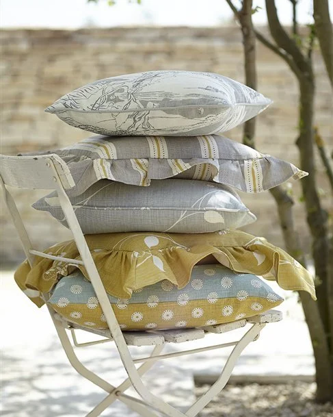 Stack of five decorative pillows on a white metal chair outdoors, with trees and a brick wall in the background.