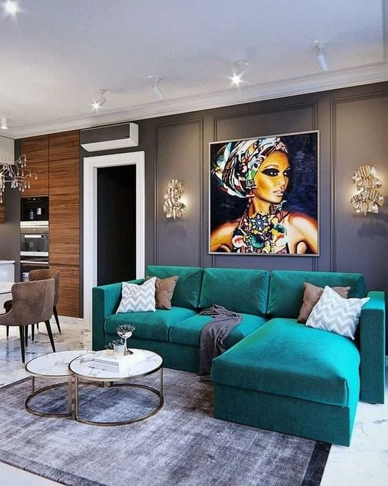 Modern living room with a teal sectional sofa, decorative pillows, and two round coffee tables on a gray rug. A colorful African art portrait hangs on a gray wall, and a dining area with a brown chair and chandelier is visible in the background.