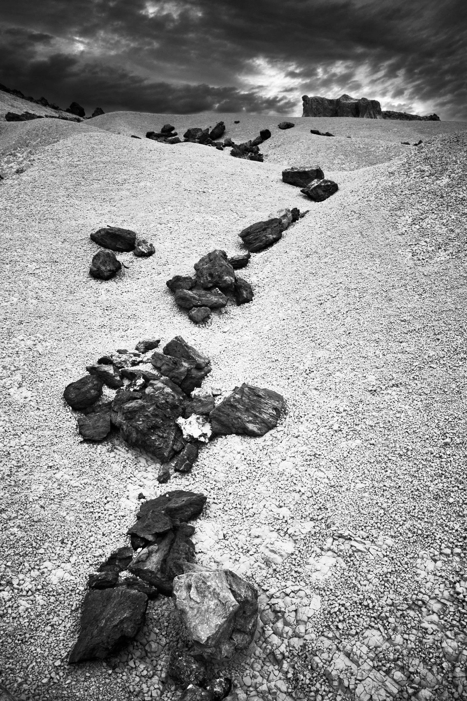 Rock Drift - A scattered run of stones moves through bright gravel like a slow current, pointing toward the higher ground and the rim beyond; Big Bend weather sits above it.