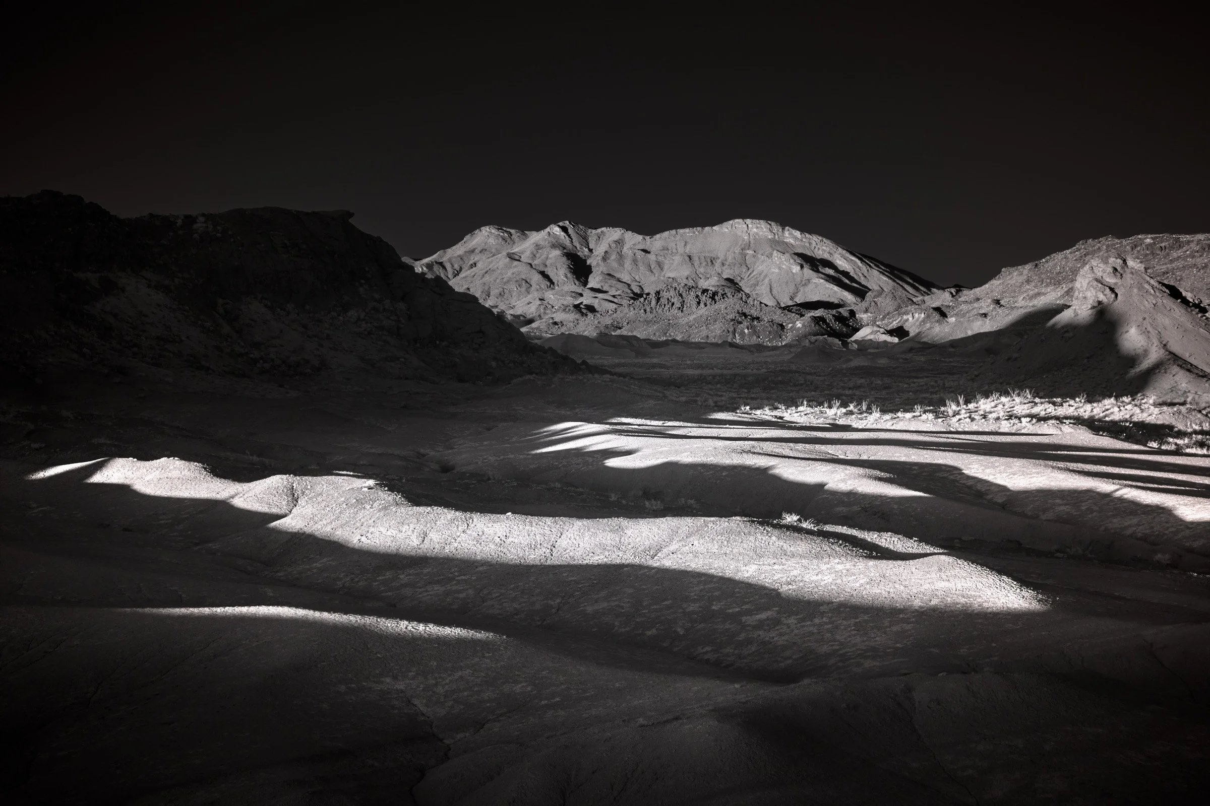 Light Strokes on Dark Basin - The ground takes light in bands and fades it back into shadow, as if the basin is being written on once and then erased; Big Bend stone stands quiet behind it.