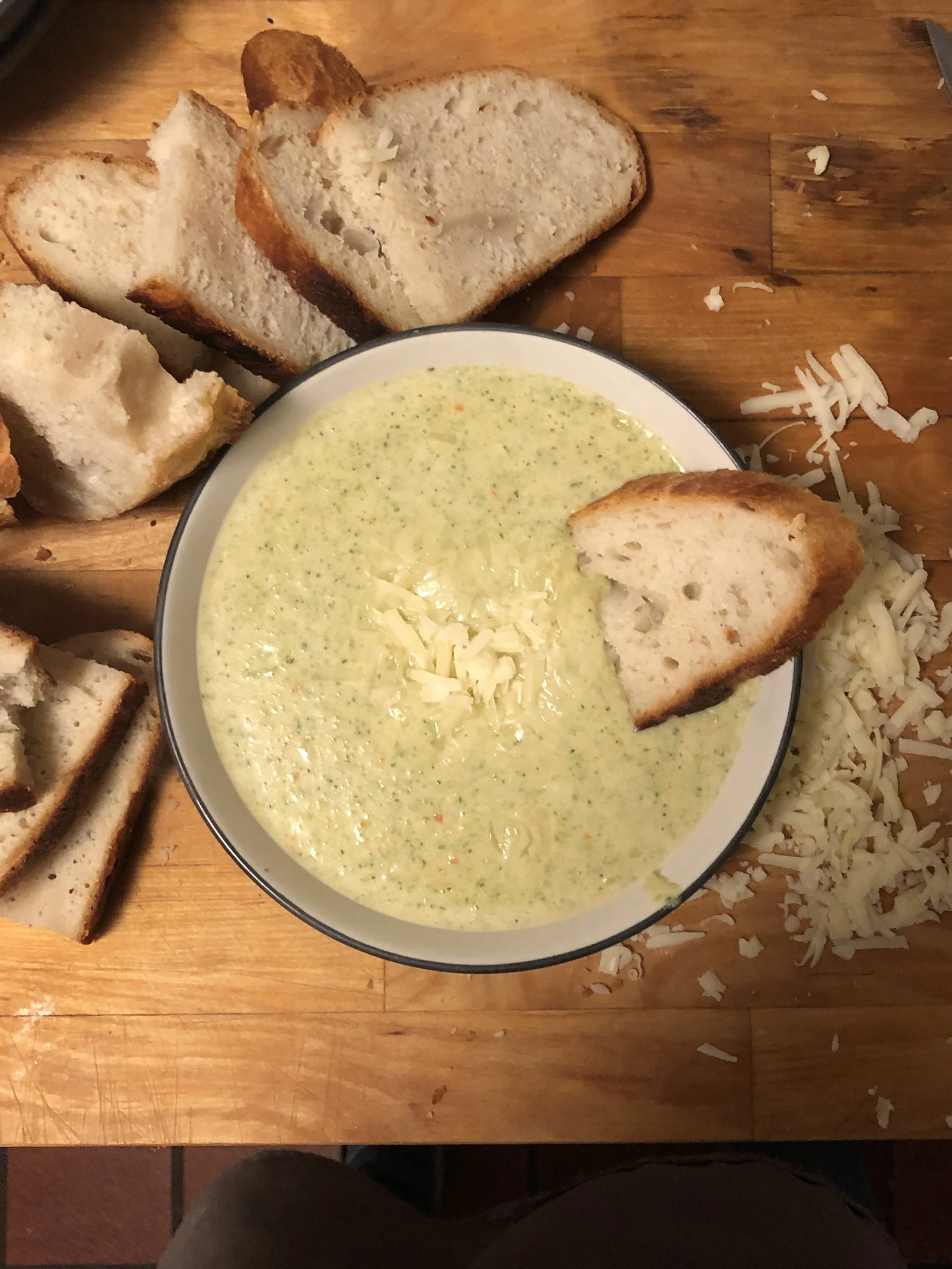 A bowl of creamy broccoli cheese soup garnished with shredded cheese, surrounded by slices of toasted bread and chunks of shredded cheese on a wooden table.
