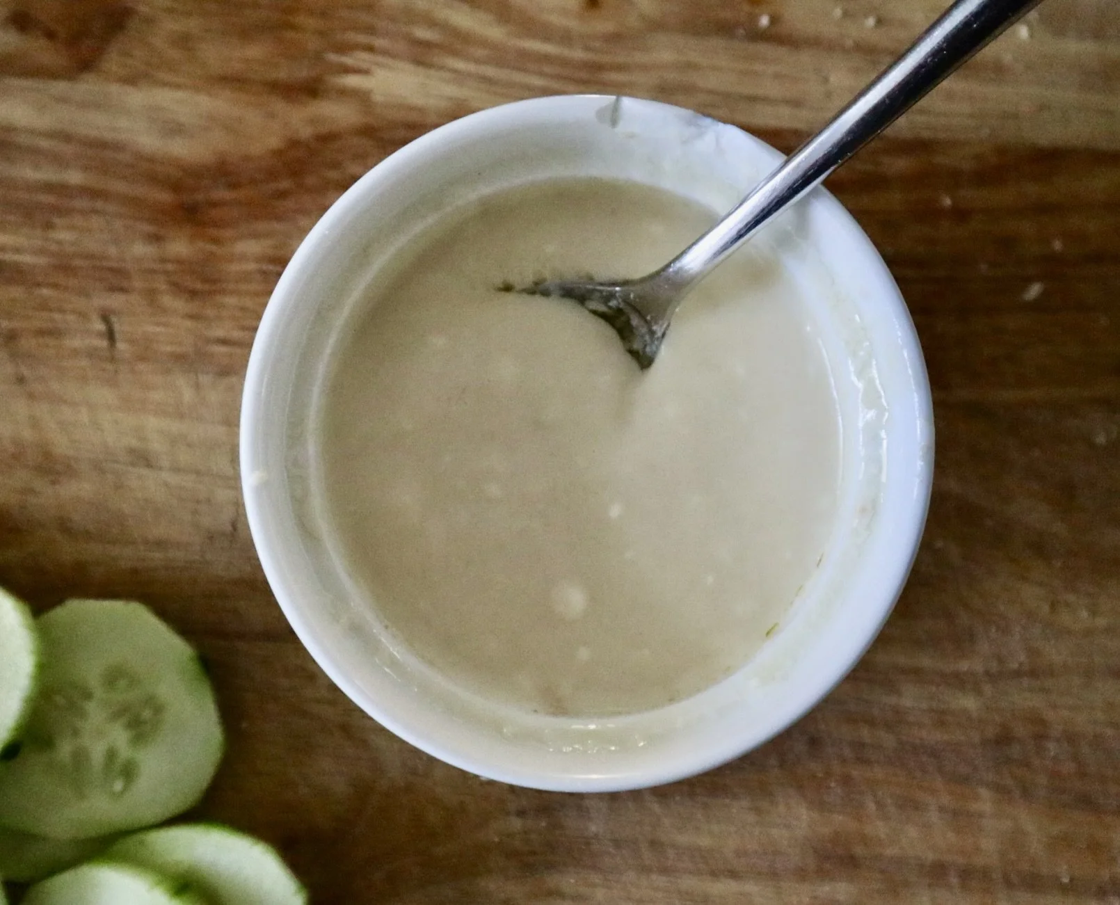 A white cup filled with a thick, creamy, light-colored sauce or dip, with a metal spoon inside, on a wooden surface with sliced cucumber on the side.
