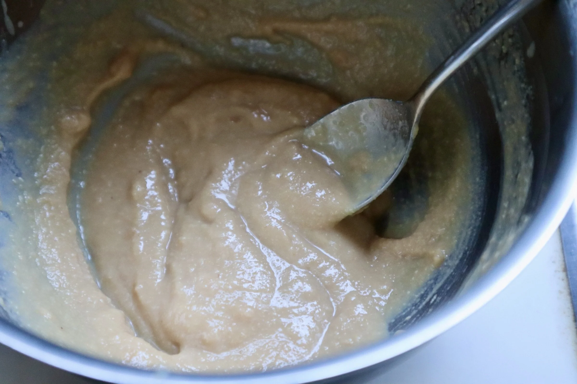 Close-up of a silver spoon stirring thick, beige-colored peanut butter in a metal container.