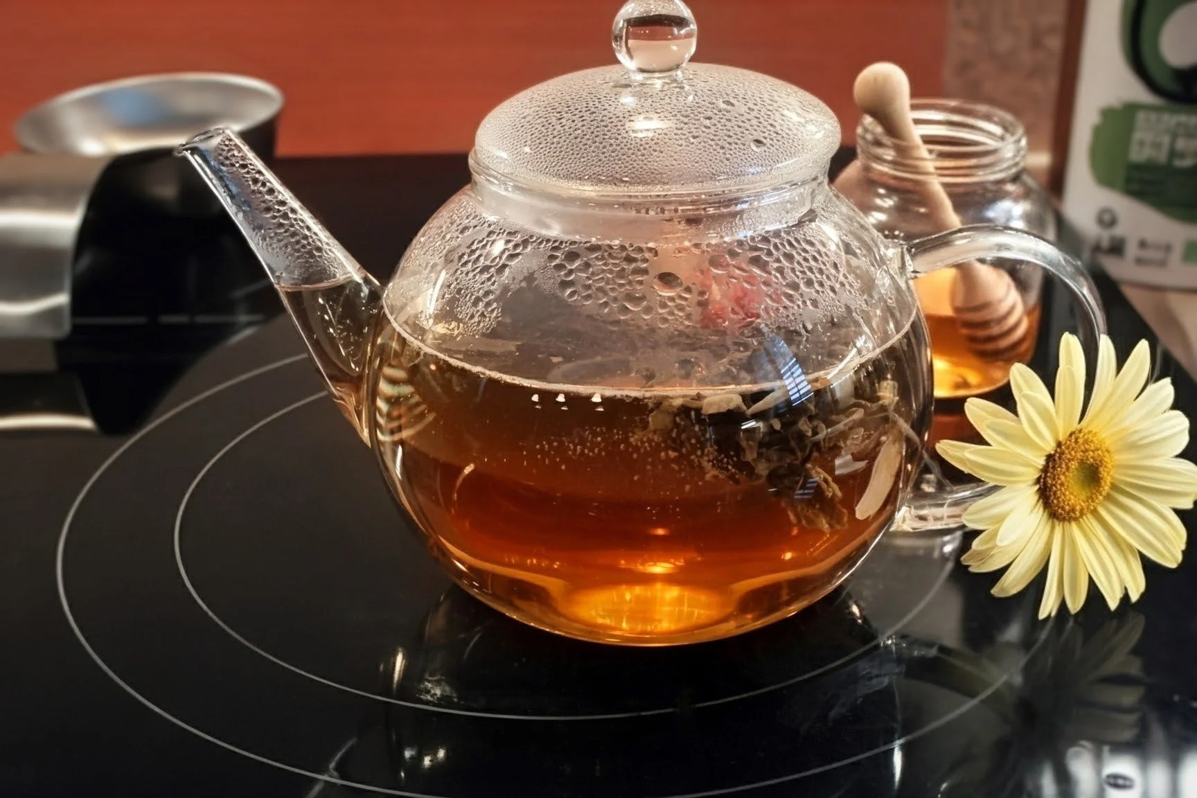 A clear tea pot with blooming flower, a honey jar, and a white flower on the stove