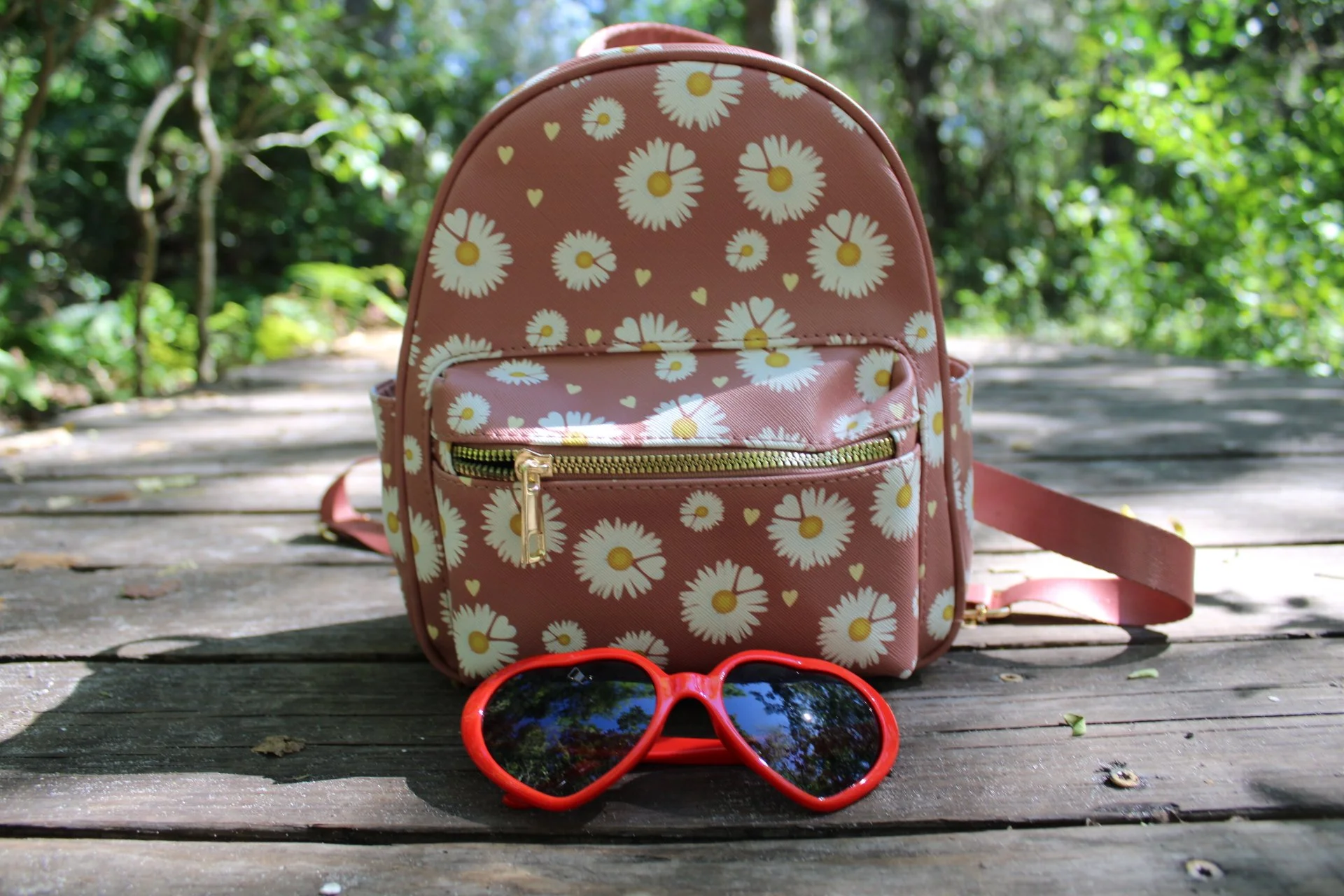 Dusty rose colored backpack with flowers for hiking plus red heart-shaped glasses in the forest