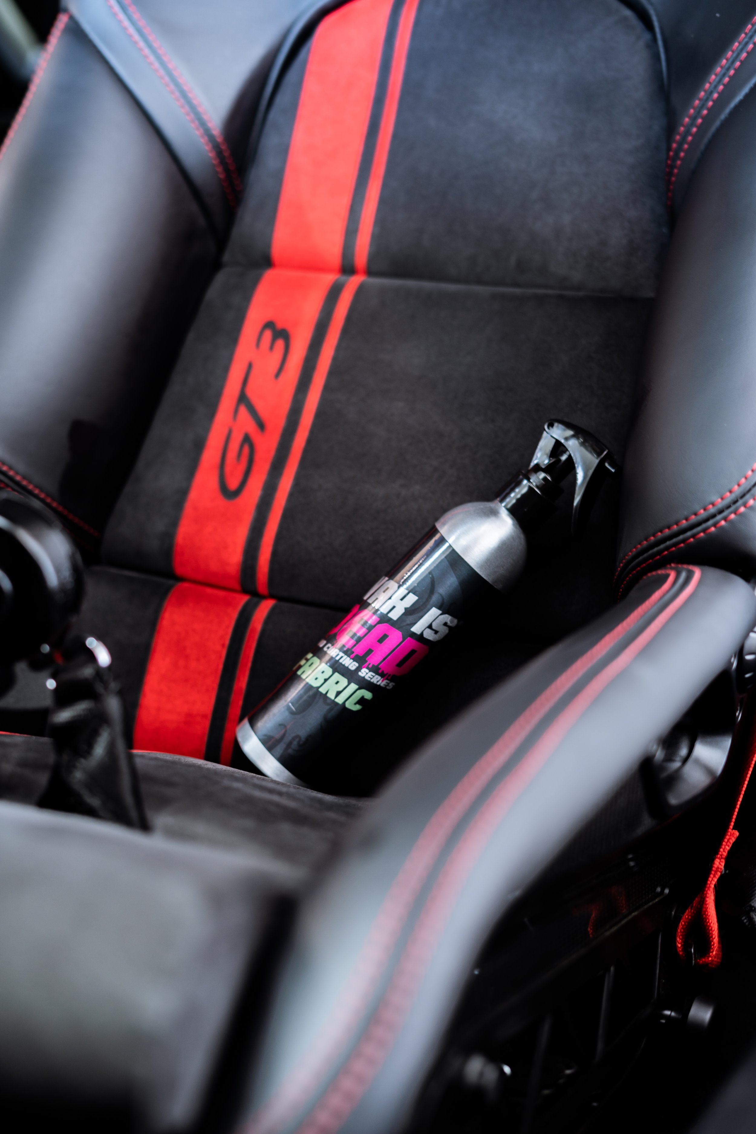 Close-up of a black and red racing car seat with the brand GTR3 printed on a red stripe, and a spray bottle resting in the seat's side pocket.