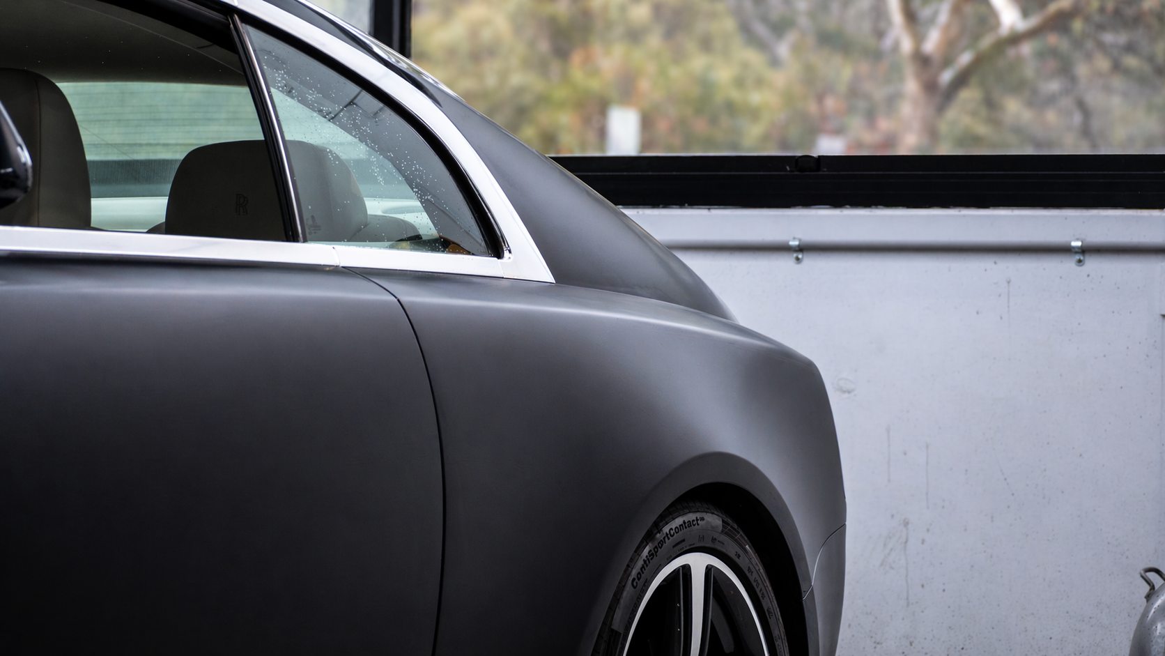 Close-up of a matte gray luxury car with a visible black wheel, parked in a concrete garage or parking area, with trees blurred in the background.
