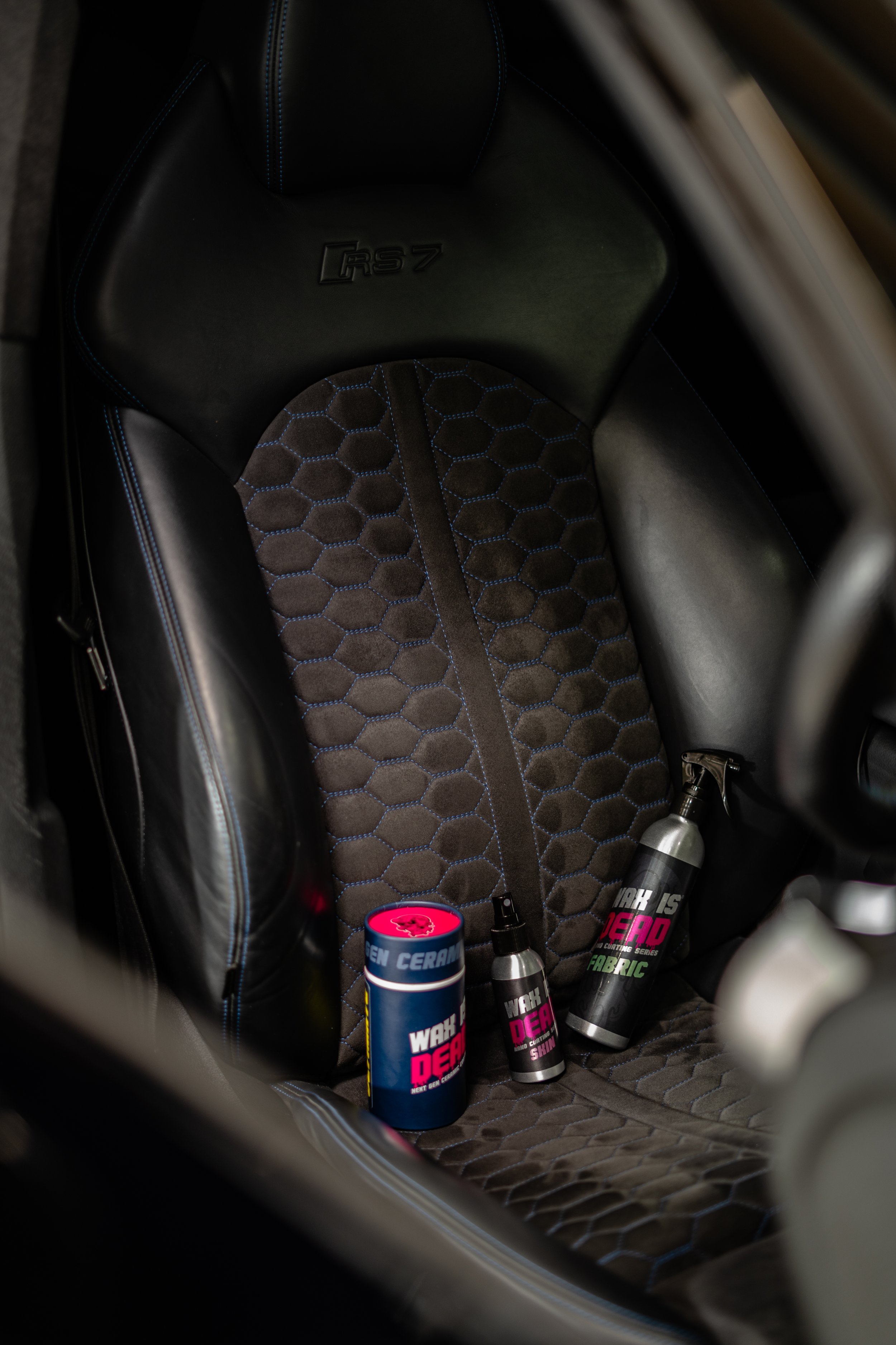 Inside view of a car seat with various cans and bottles of detailing or cleaning products, including a can labeled 'WAX DEAR' and a bottle labeled 'FABRIC', situated on the seat's surface.
