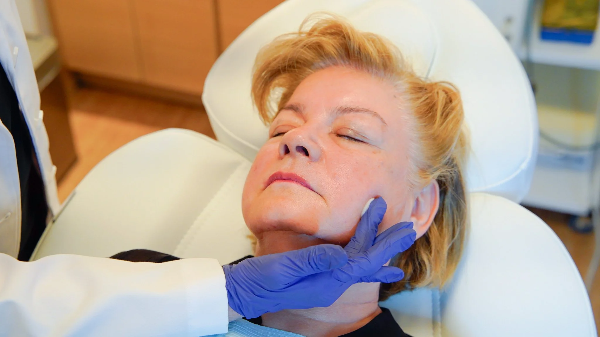 An older woman with closed eyes lying on a medical examination chair, with a healthcare professional wearing blue gloves gently touching her jawline.