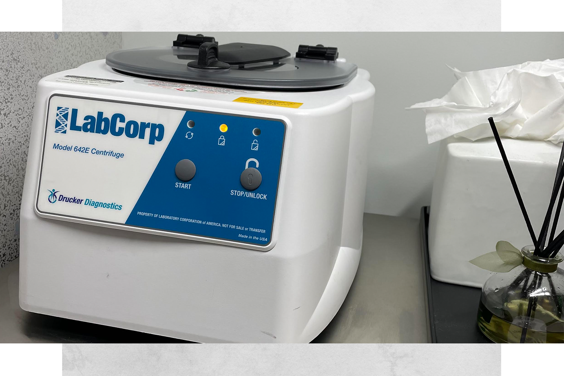 A LabCorp Model 642E centrifuge in a laboratory setting on a counter with a tissue box and a small glass vase with black sticks and a decorative ribbon.