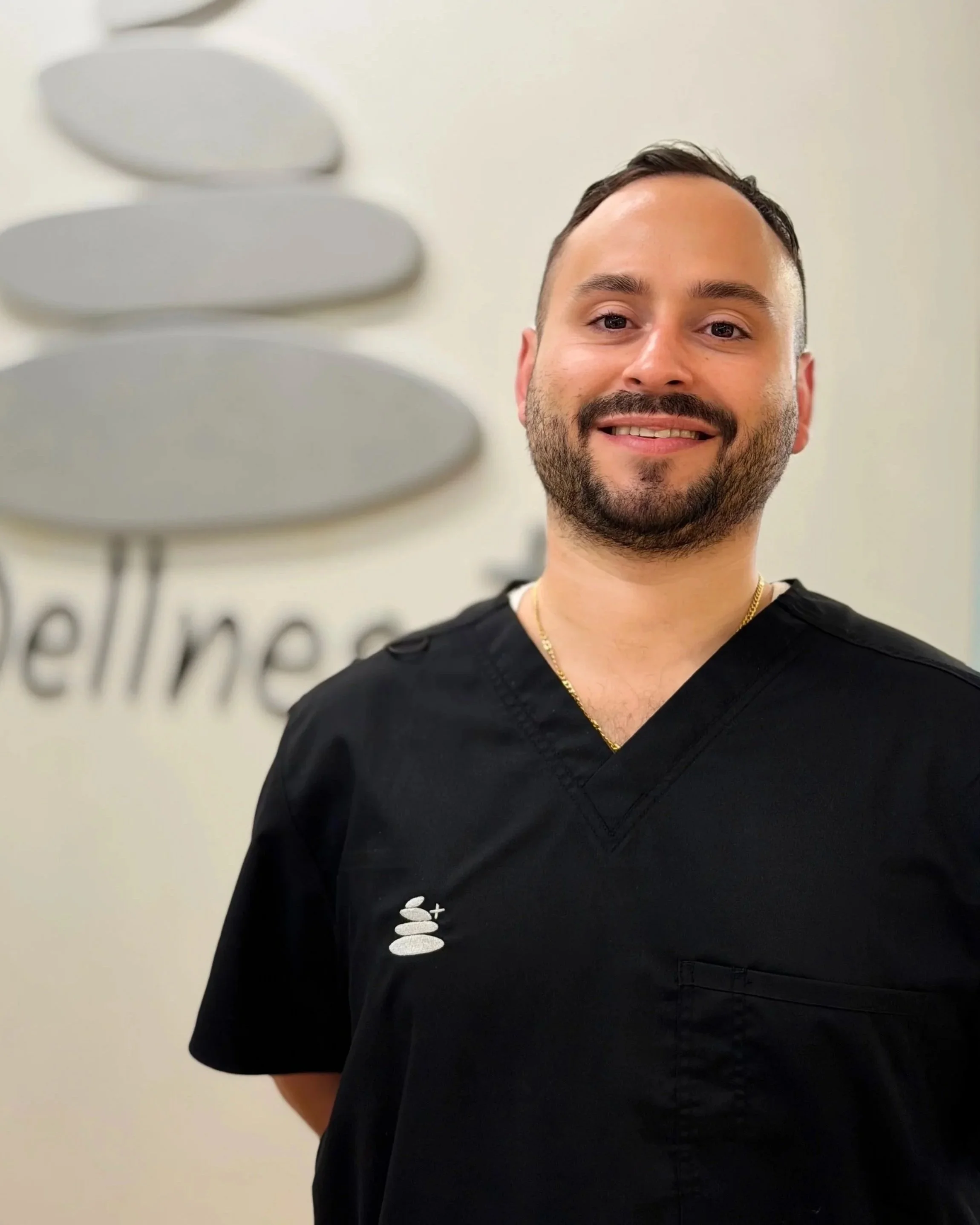 A smiling man wearing black scrubs standing in front of a wall with a logo that includes stylized stones and the word 'wellness'.