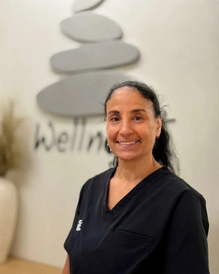A woman with dark hair tied back, wearing black medical scrubs, smiling in front of a wall with a gray abstract art piece and the word 'Wellness' partially visible.