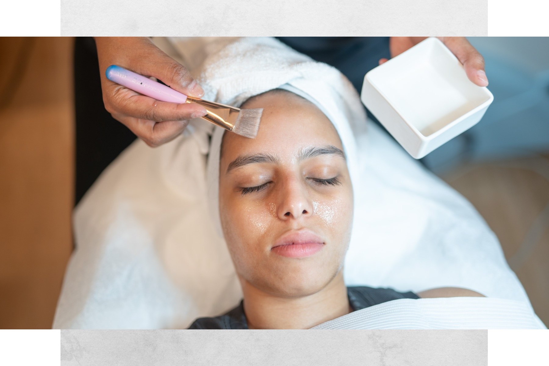 A woman receiving a facial treatment while lying down with her eyes closed, a towel wrapped around her head, and her face covered in a facial mask. An esthetician applies skincare with a brush, holding a small white container.