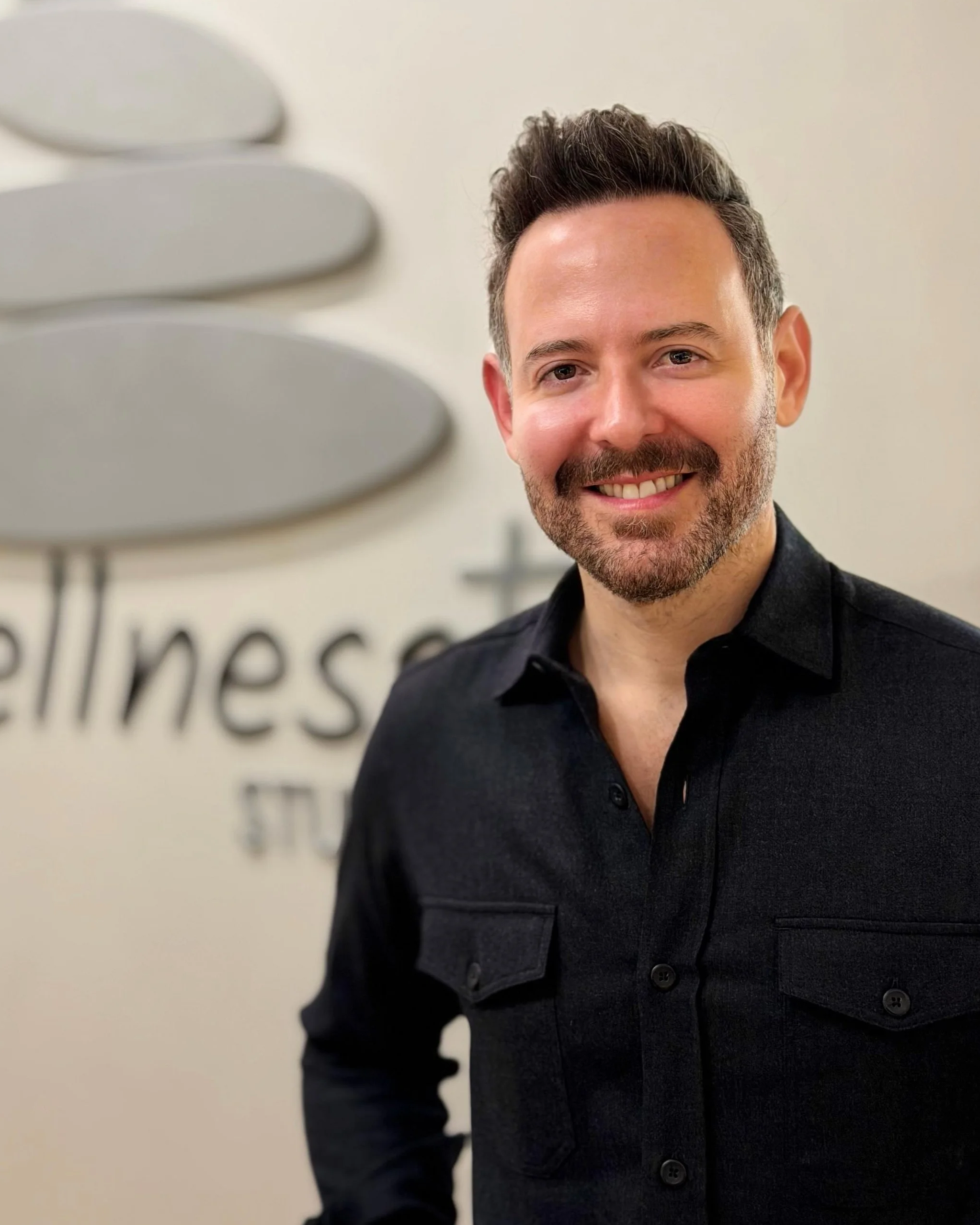 A smiling man with short dark hair and a beard, dressed in a black shirt, stands in front of a wall with a logo that says 'wellness'.
