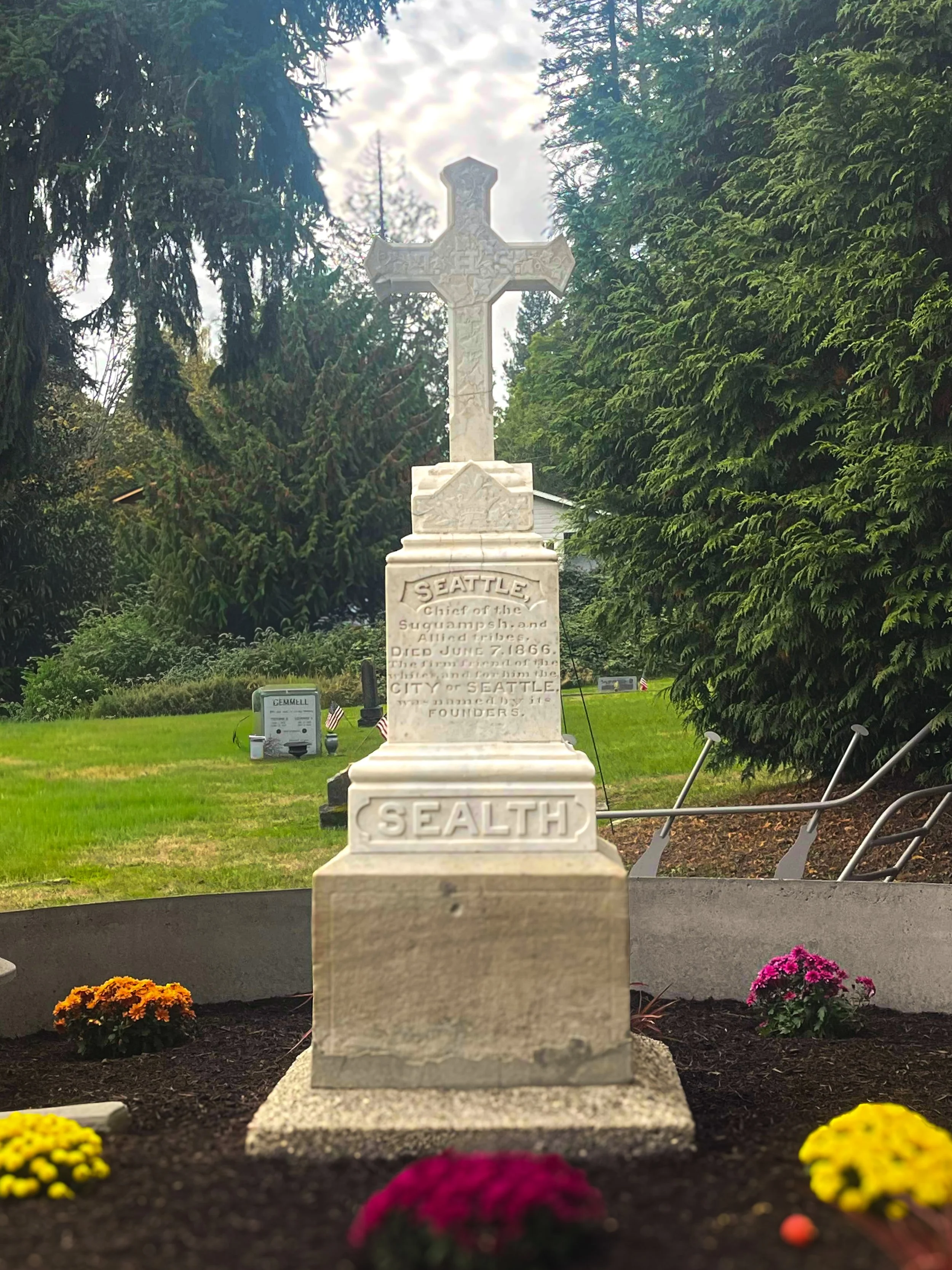 A white gravestone with a cross on top, surrounded by flowers, trees, and a green lawn.