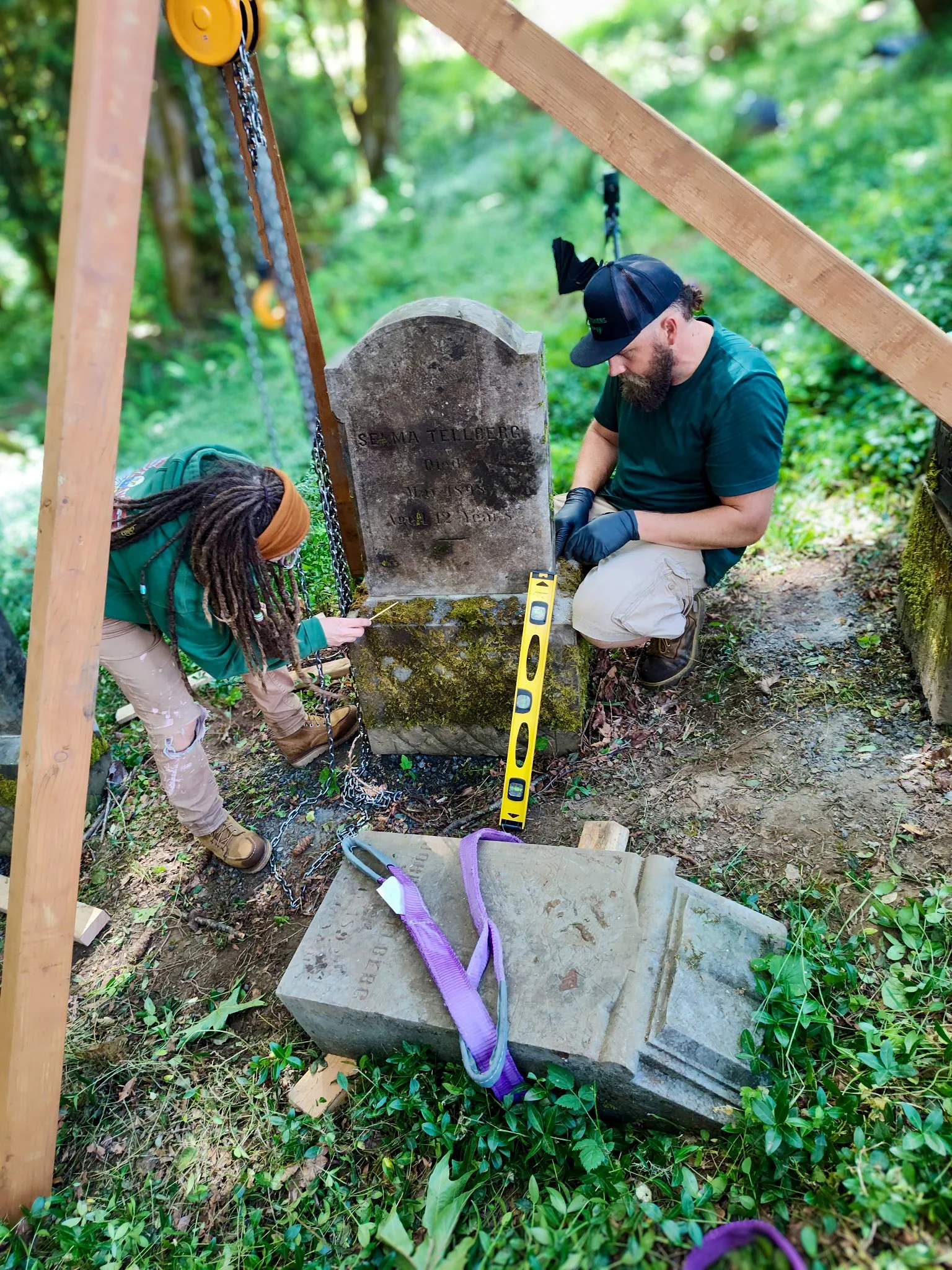 Two people working on a historic headstone outdoors. One woman with dreadlocks and an orange headband is measuring or inspecting the stone. One man with a beard and baseball cap is kneeling next to the headstone, possibly working on or examining it. The headstone has moss and weathering, with a loose broken piece of the stone next to it.