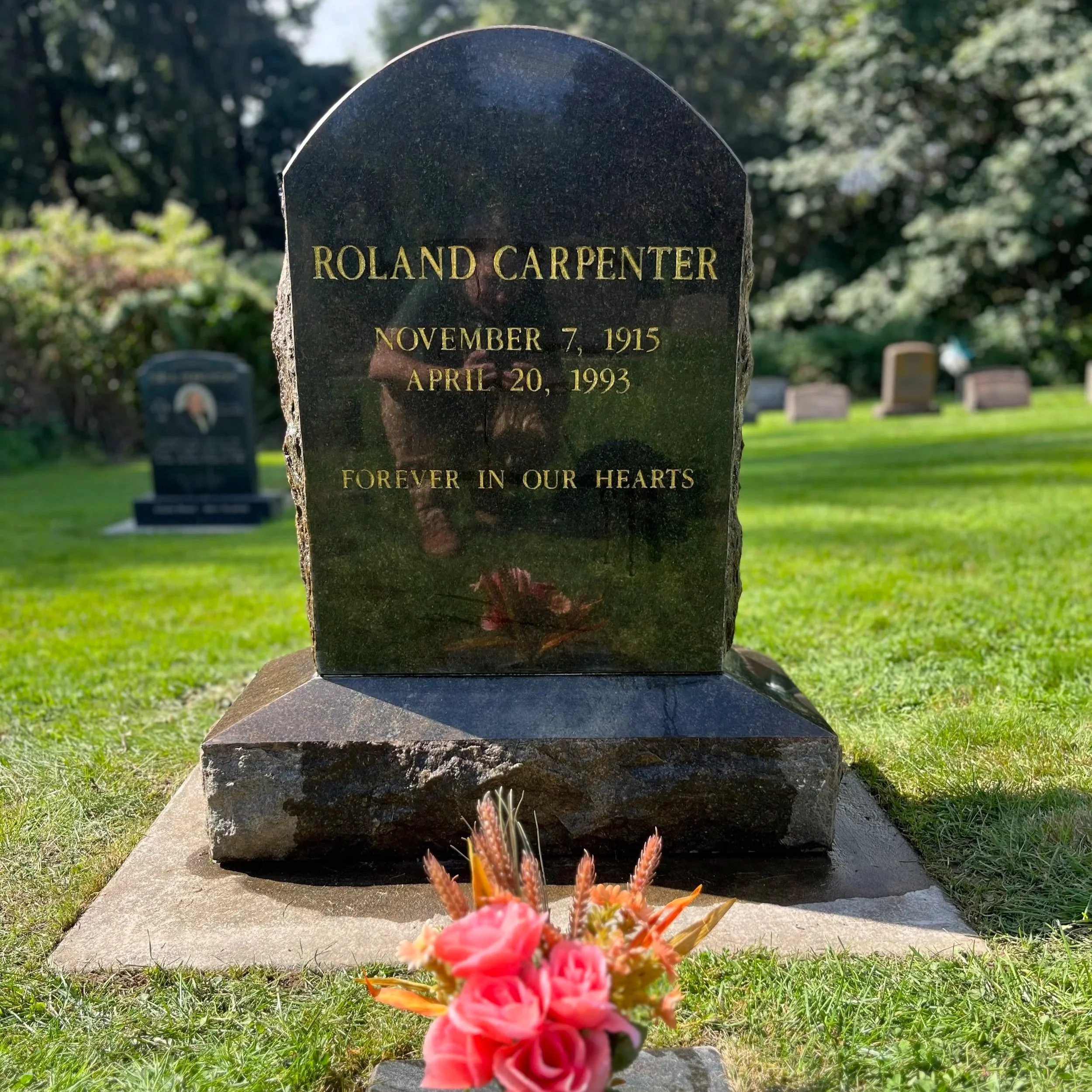 Headstone for Roland Carpenter in a cemetery, with flowers in front, surrounded by grass and other headstones in the background.