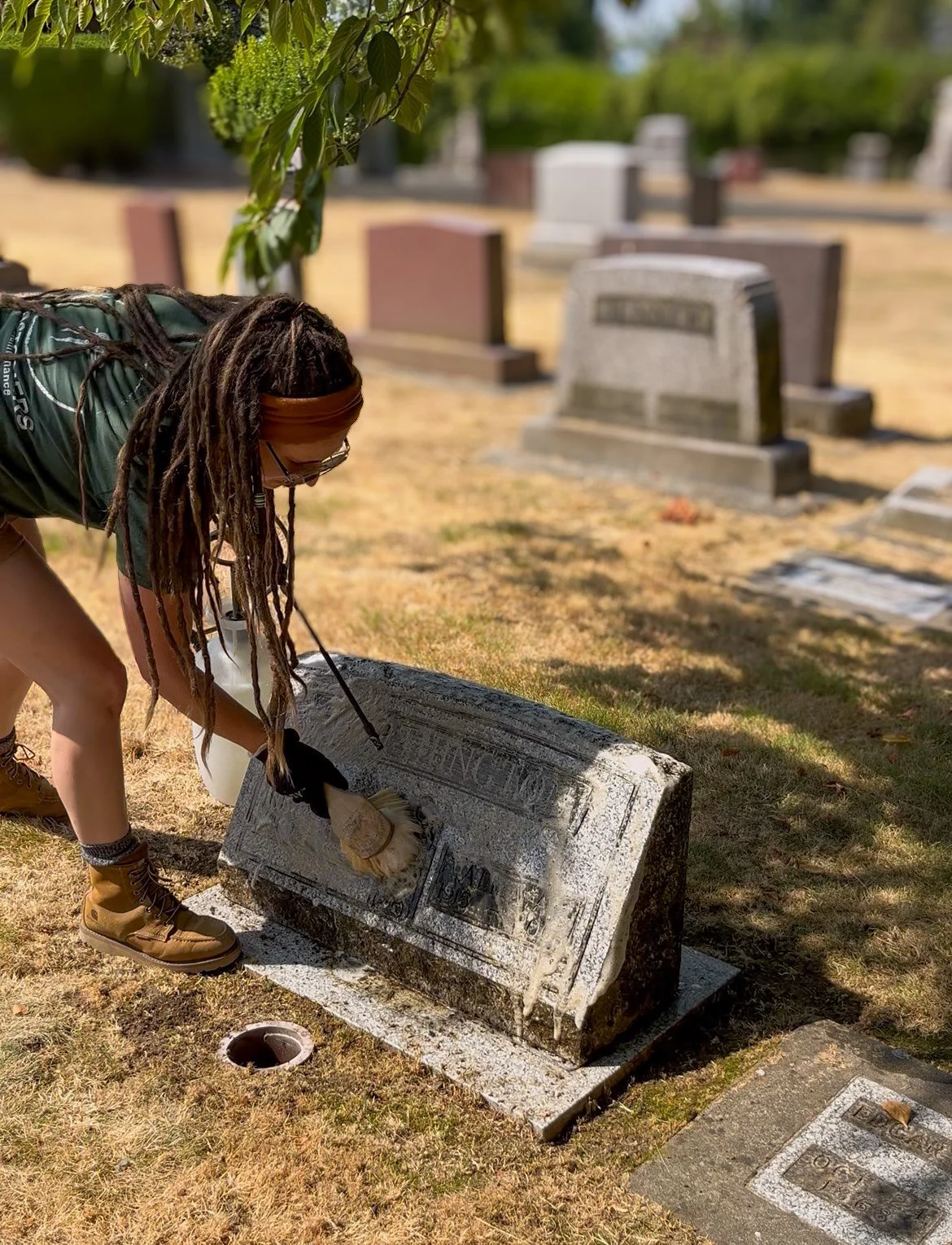 Person cleaning a gravestone in a cemetery on a sunny day.