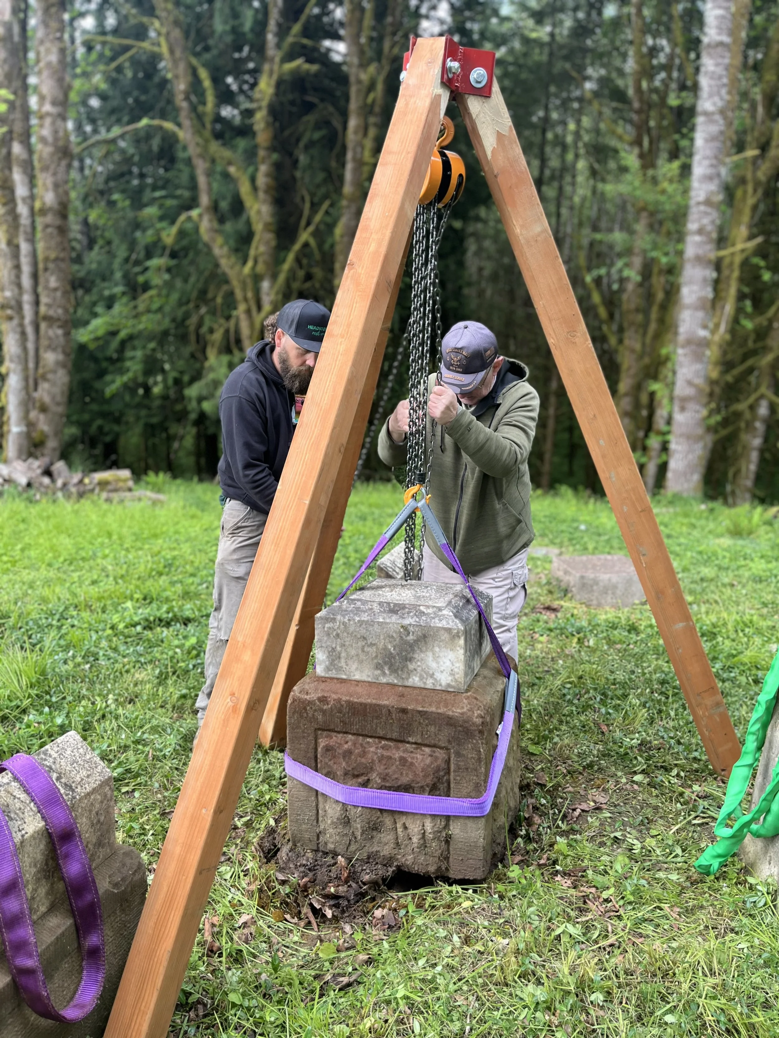 Two men lifting large headstone using a pulley system in a grassy, wooded area.