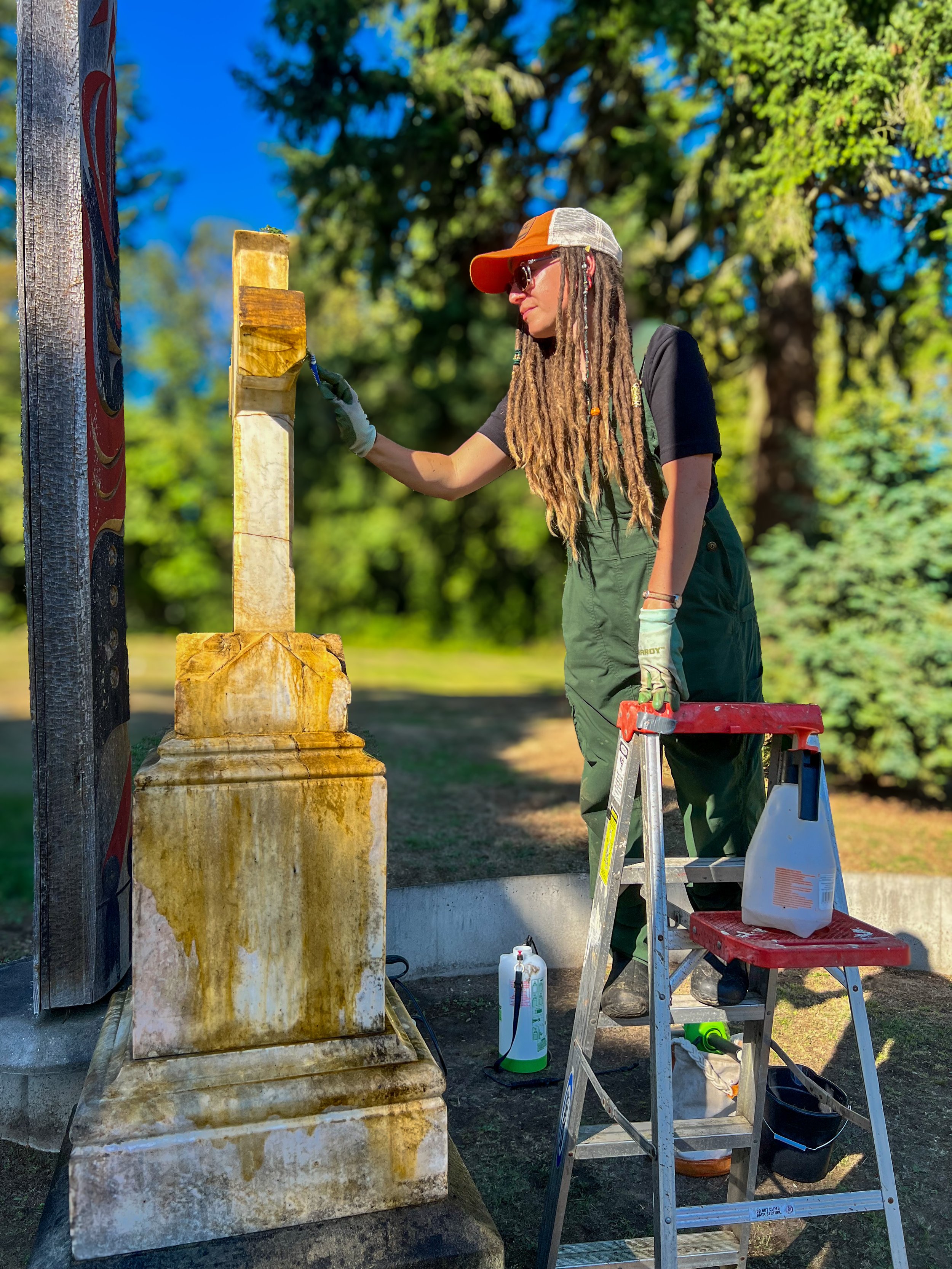 A woman with long dreadlocks and wearing a hat and sunglasses is cleaning or restoring a historic stone monument using a brush. She is standing on a ladder, outdoors, with trees and a bright blue sky in the background.