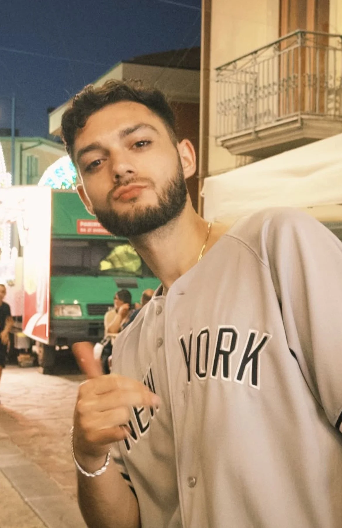 Alt text: Mikey Fiume in a New York Yankees jersey standing on an Italian village street at dusk — New York raised, Italian at the core.