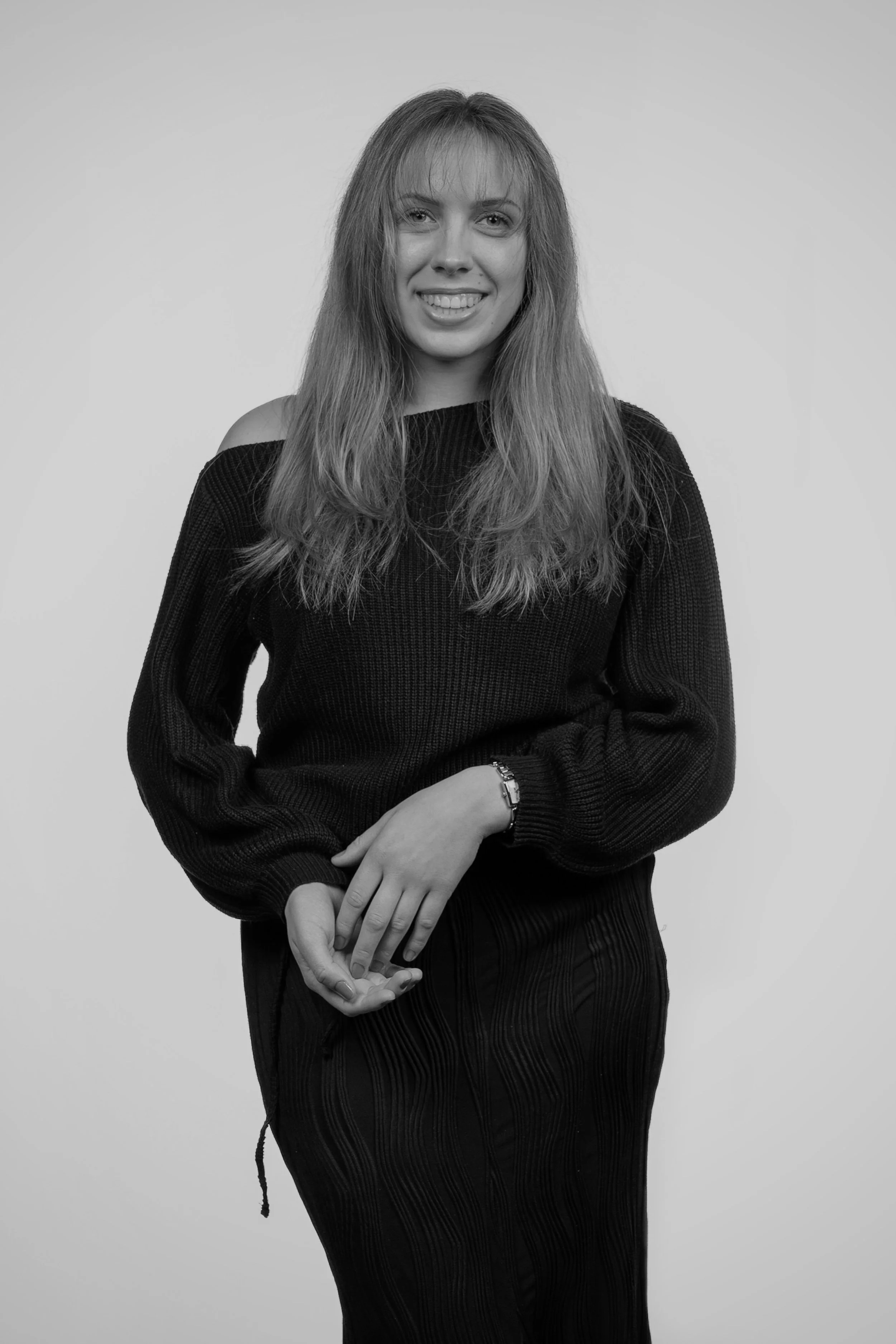 Black and white portrait of a young woman with long hair, smiling, wearing a dark sweater and skirt, standing against a plain background.