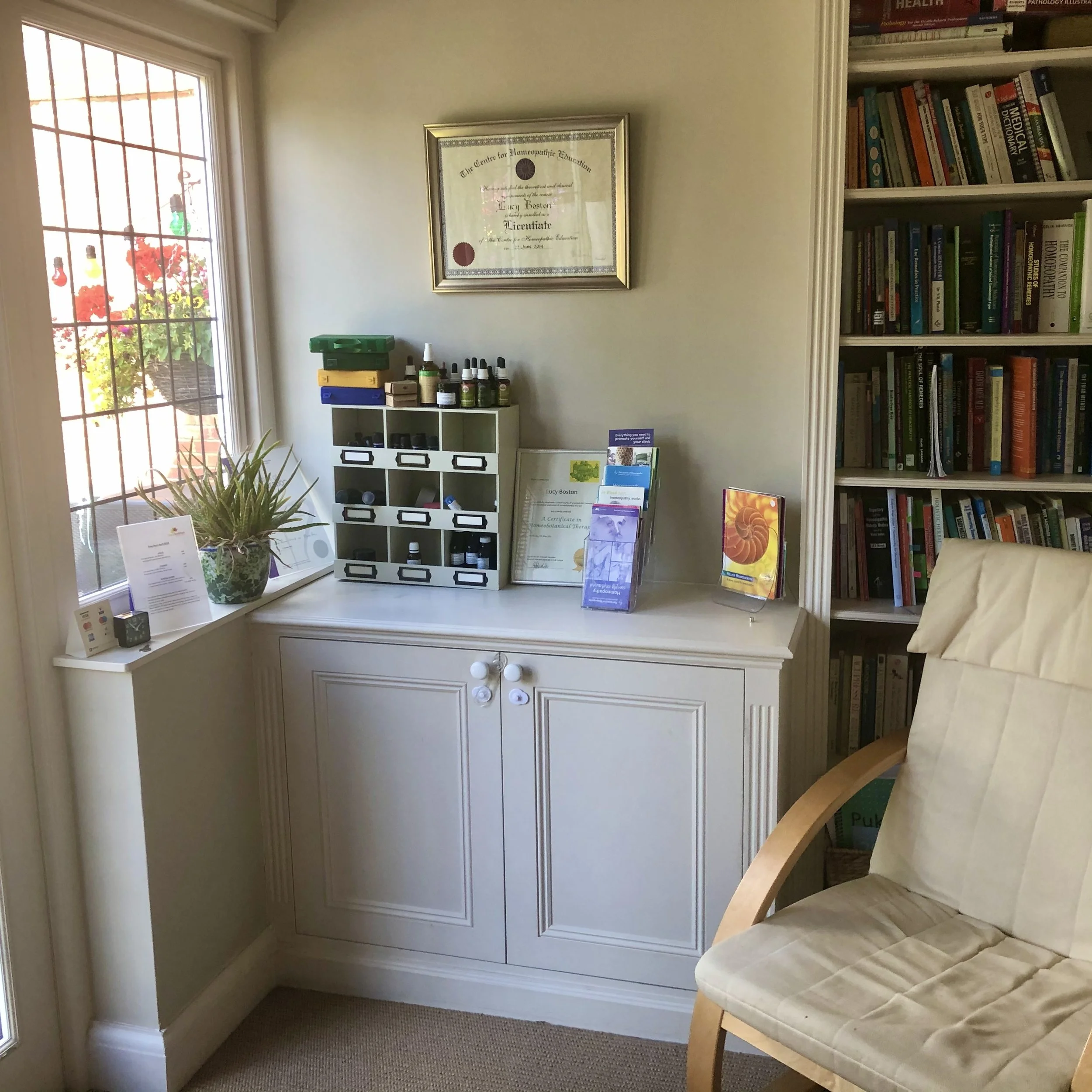 Corner of a room with a white cabinet, a bookshelf filled with books, and a cream-colored chair. On the cabinet, there are essential oils, brochures, and a framed certificate hanging on the wall. A window with bars and a potted plant on the windowsill are visible, with bright natural light coming in.