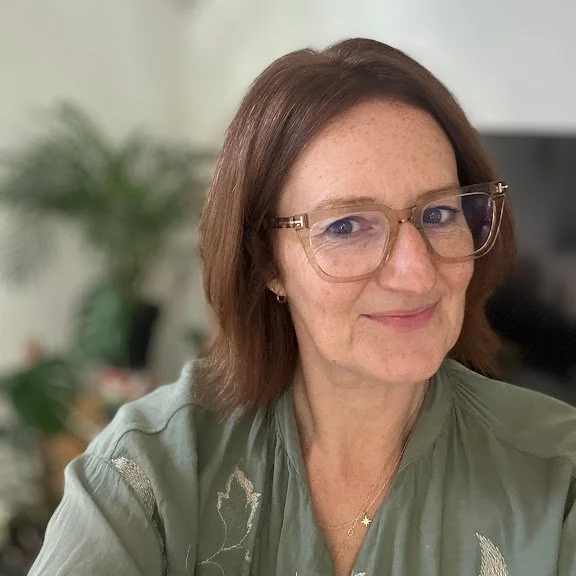 A woman with shoulder-length brown hair and glasses, smiling in a well-lit room with a plant in the background.