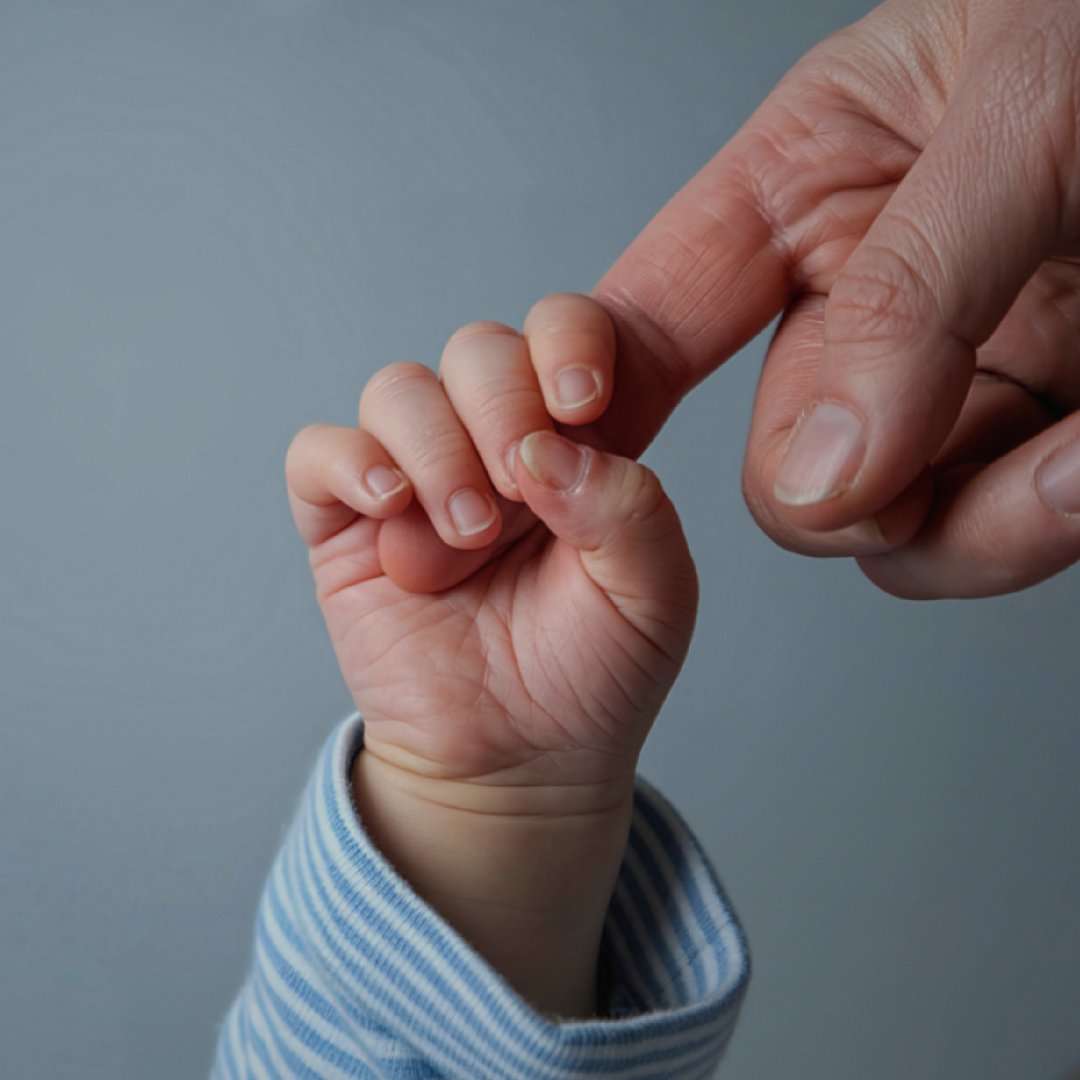 An infant hand wraps around an adult finger
