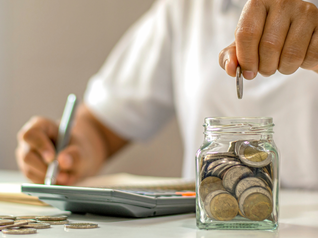 person in white shirt drops coins into a jar, sitting in front  of a calculator and pen