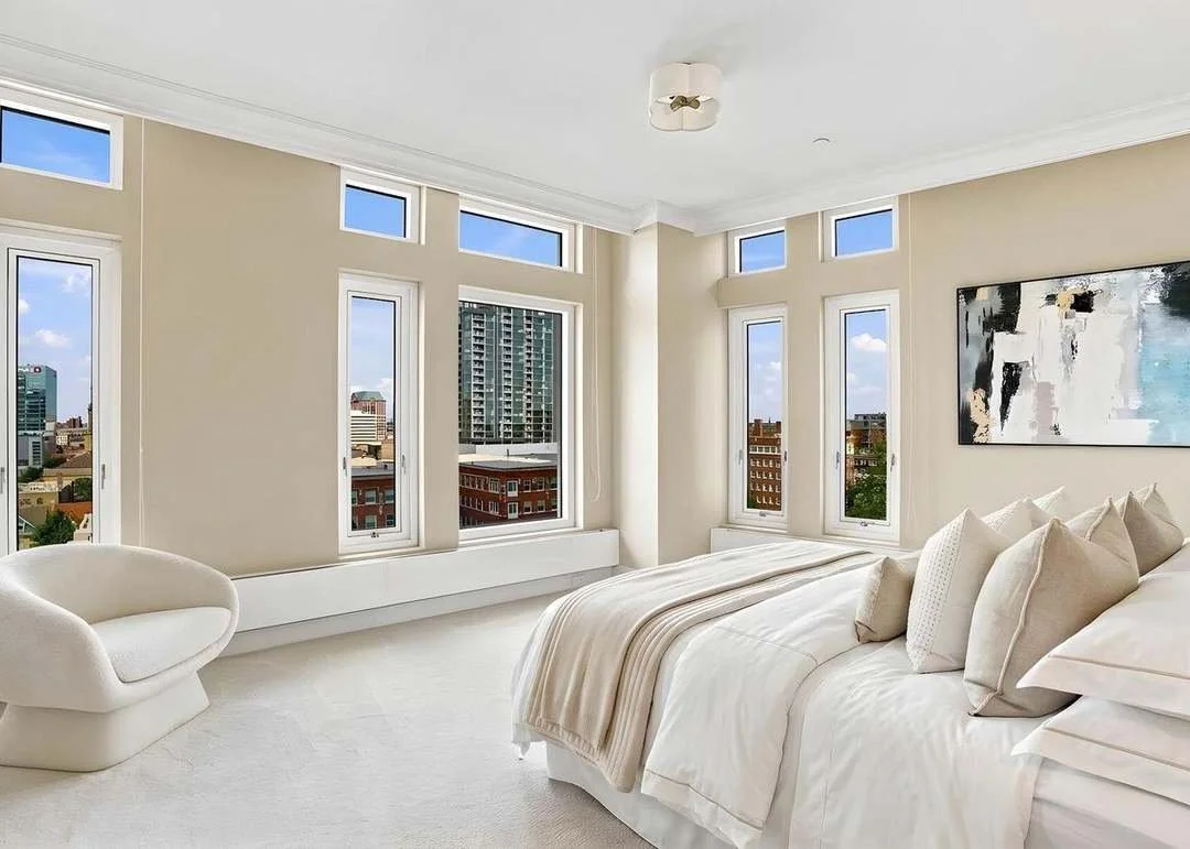 Bright bedroom with large windows, a white bed with beige pillows and blanket, a modern white chair, and a city view outside.