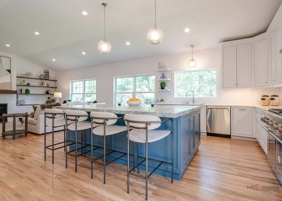 Bright open-concept kitchen with white cabinets, a large blue island with a marble countertop, and white bar stools. Light wooden floors, large windows, and minimal decor with built-in shelves and a cozy living area in the background.