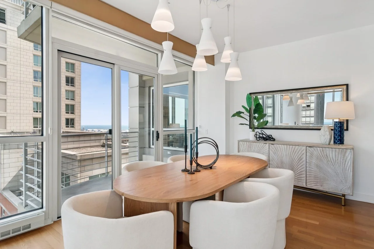 Modern dining area with round wooden table, six white upholstered chairs, large sliding glass doors leading to a balcony, and city and ocean view outside. Decor includes a long sideboard with a large mirror, potted plant, chandelier with multiple white pendant lights, and a lamp with a white shade and blue vase.