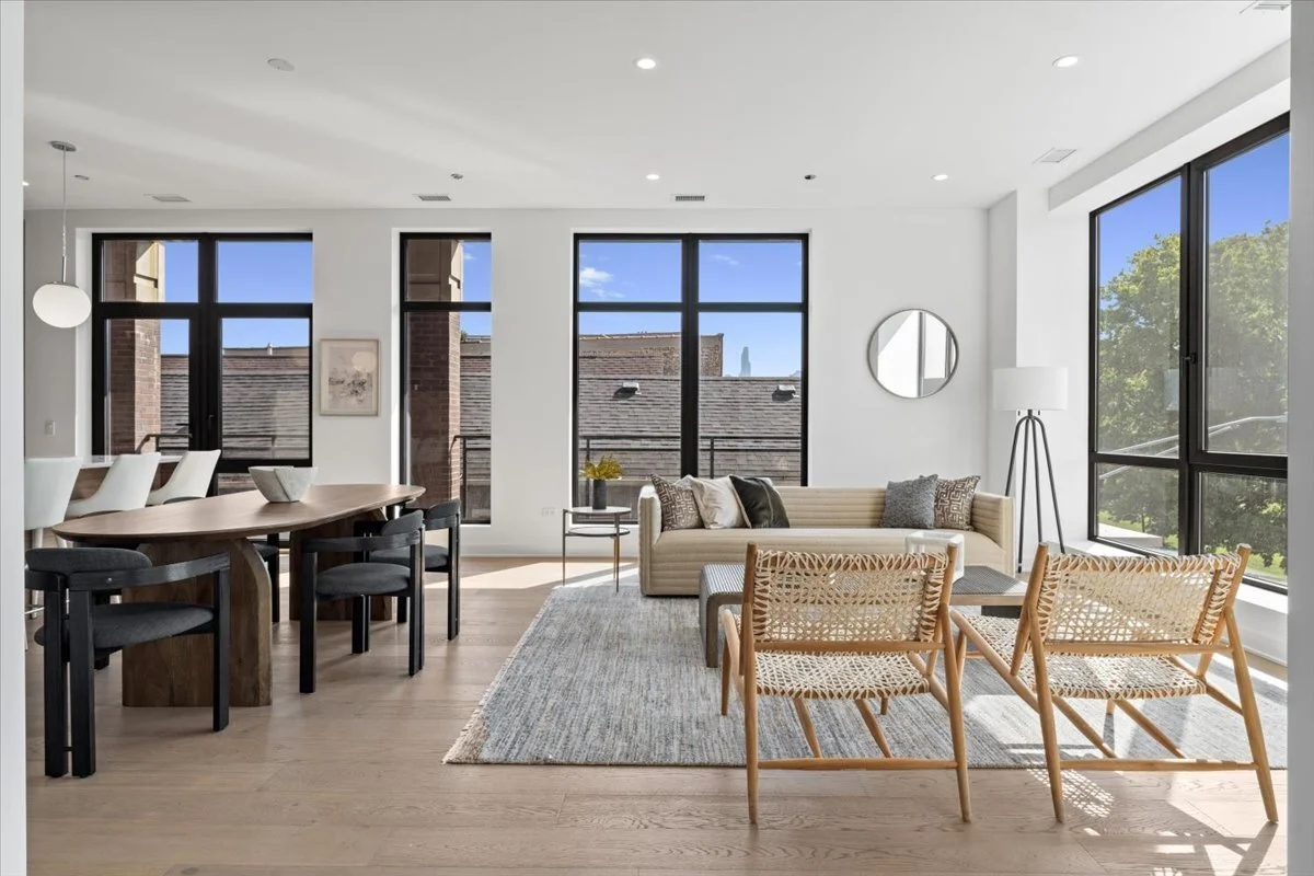 Bright, modern living room with large windows, a beige sofa, two woven chairs, a gray area rug, a white floor lamp, and a dining table with black chairs.