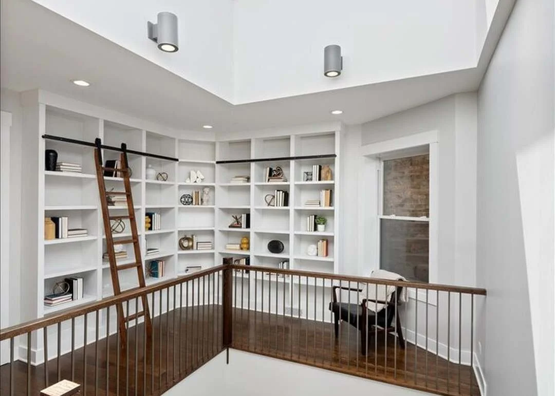A corner of a modern room with white bookshelves filled with books and decorative objects. There is a wooden ladder leaning against the shelves, a small black chair, and a skylight ceiling with recessed lighting.