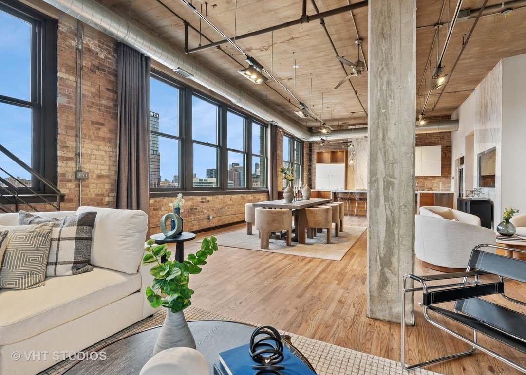 Open-concept loft apartment with large windows, exposed brick walls, wood flooring, modern furniture, a dining area with a round table and chairs, and a kitchen in the background.