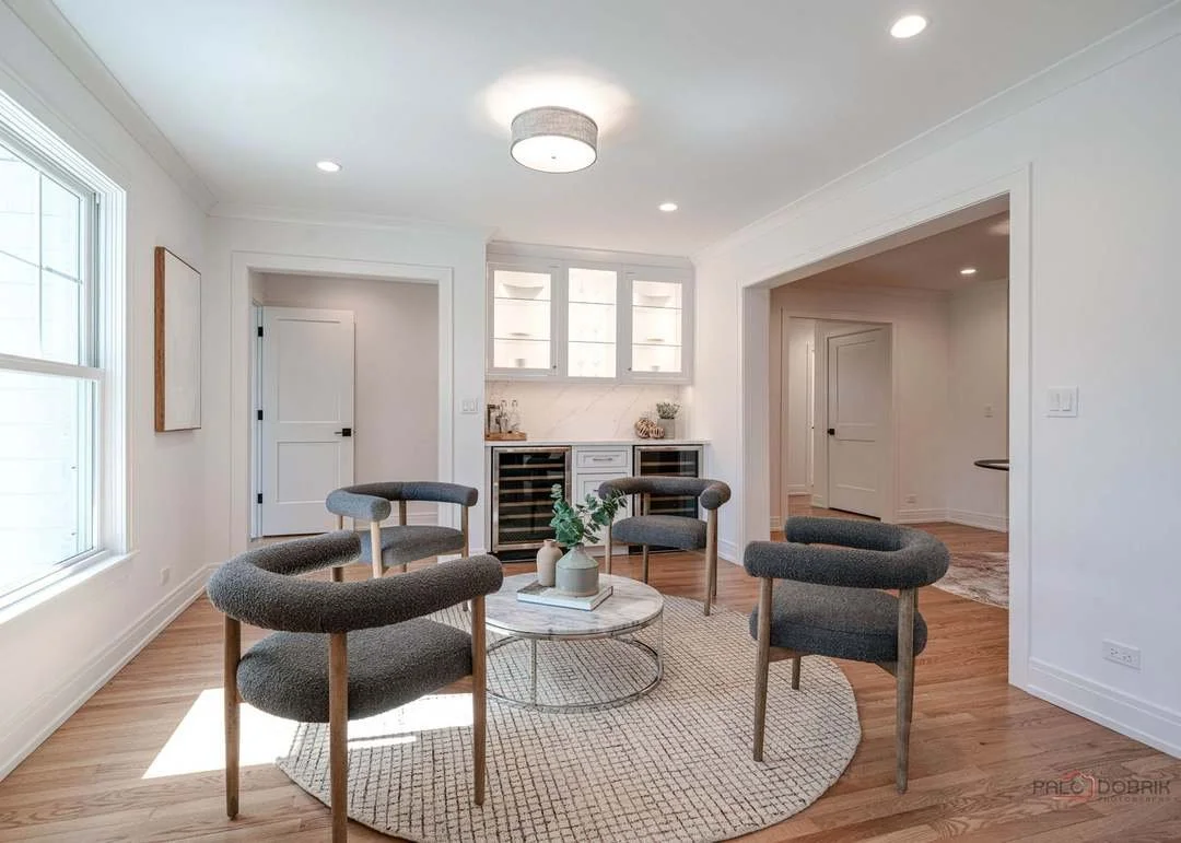 Modern living room with four gray upholstered chairs around a round marble coffee table, a built-in bar area with glass-front cabinets and wine refrigerators, white walls, hardwood floors, large window, and ceiling light.
