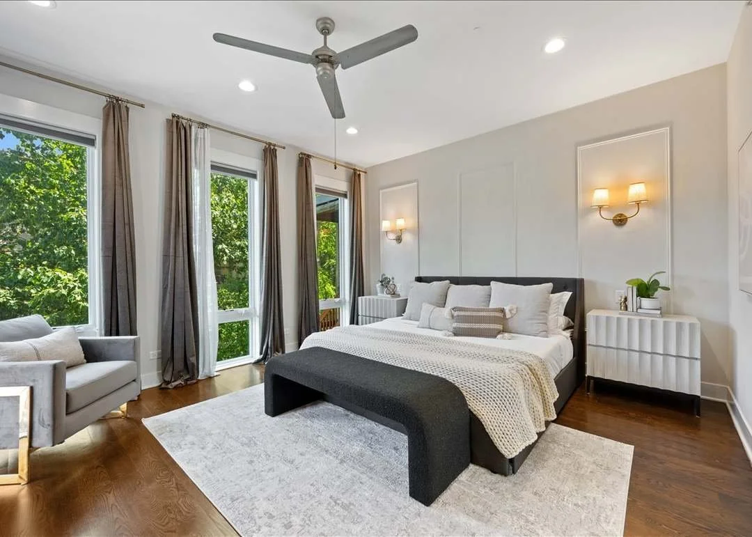 Modern bedroom with large windows, beige curtains, a bed with white bedding, a gray bench at the foot of the bed, nightstands with lamps, an armchair, and a white ceiling fan.