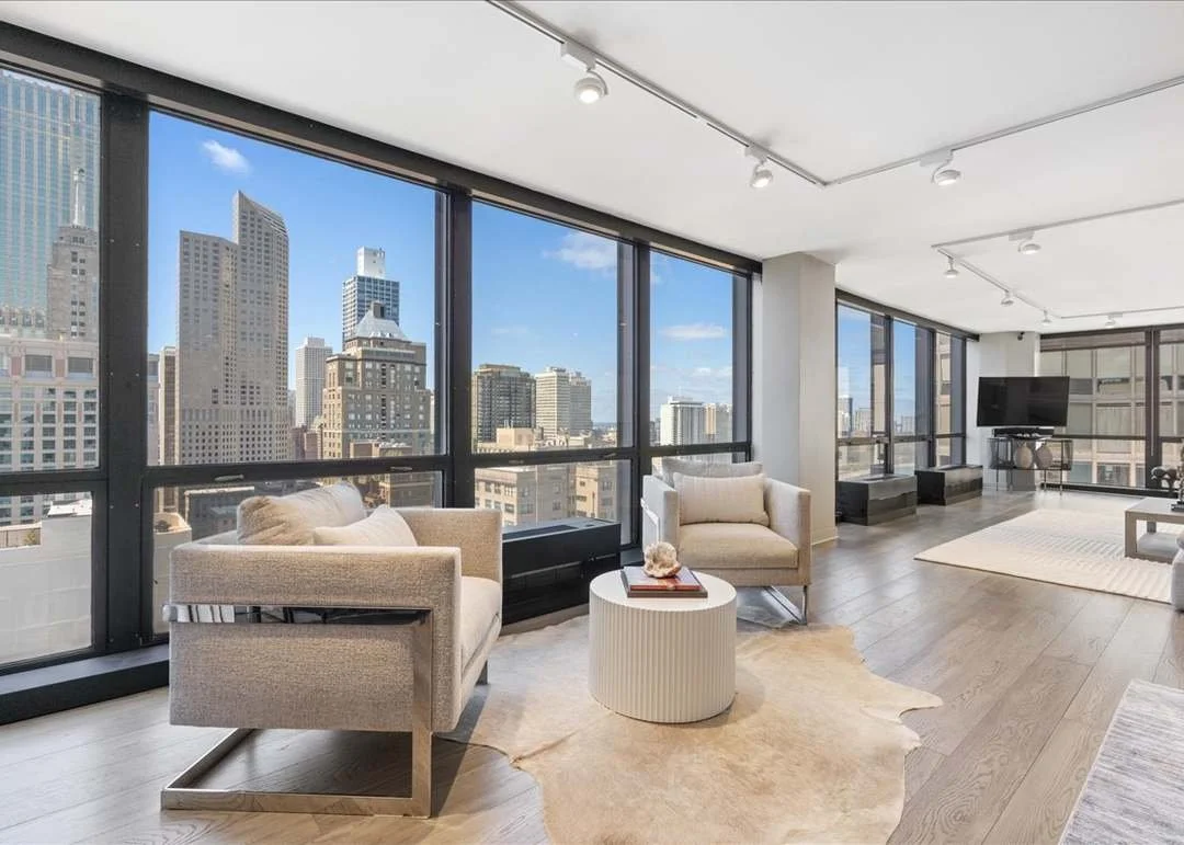 Modern living room with large windows overlooking a city skyline, featuring beige sofas, a round coffee table with decorative items, a television, and a light-colored rug on wooden flooring.