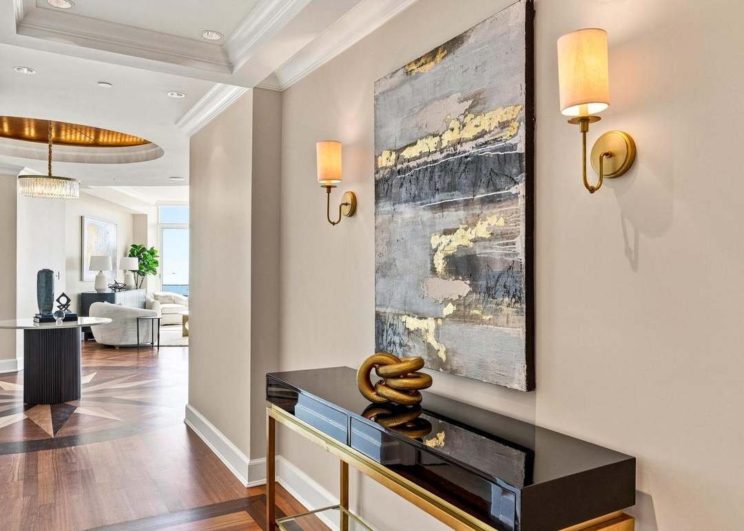Modern living room interior with wall-mounted lights, abstract art, a decorative gold sculpture on a black console table, and a glimpse of a sitting area with white furniture and a window.